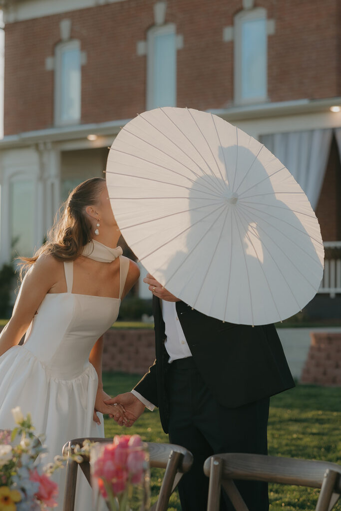 Bride and groom kissing behind a white parasol in front of a historic manor
