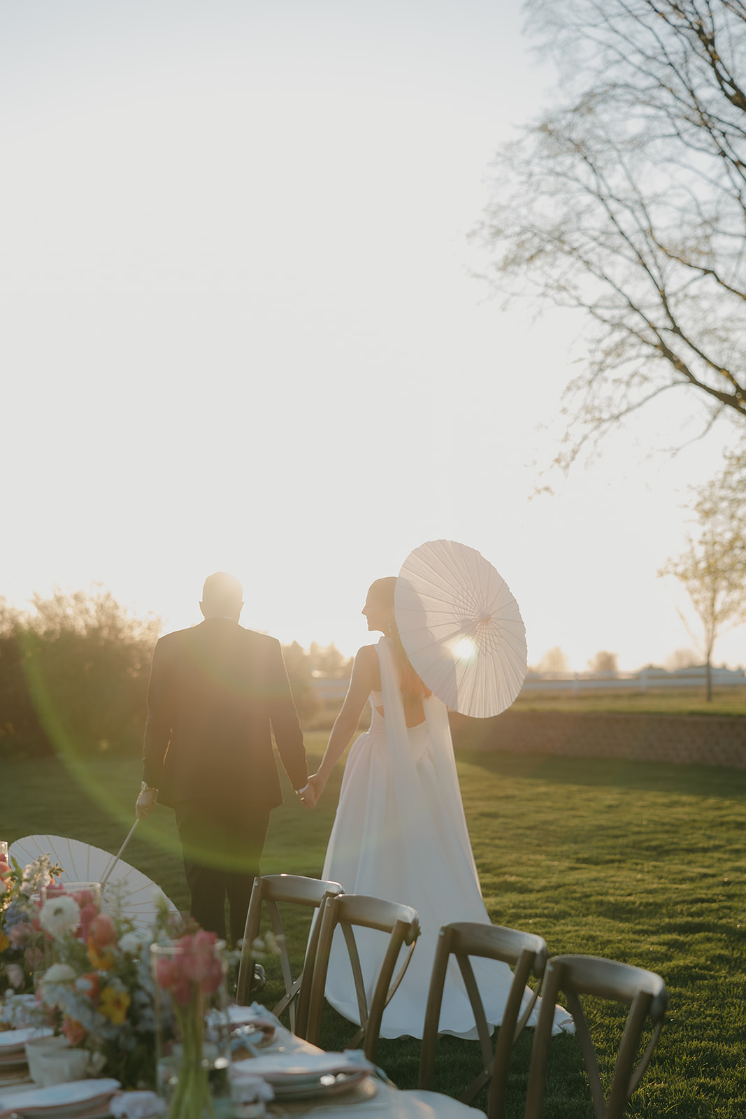 Bride with a parasol and groom walking hand in hand past their floral reception table into a glowing old money wedding sunset, the kind of moment you never want to end
