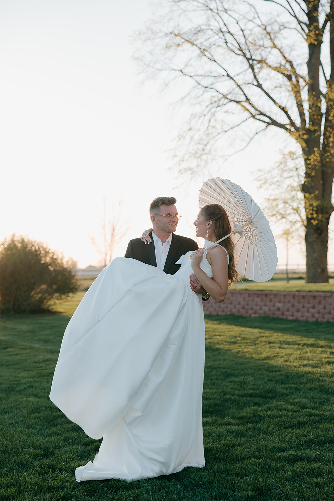 Groom lifting bride in a white gown with parasol at sunset
