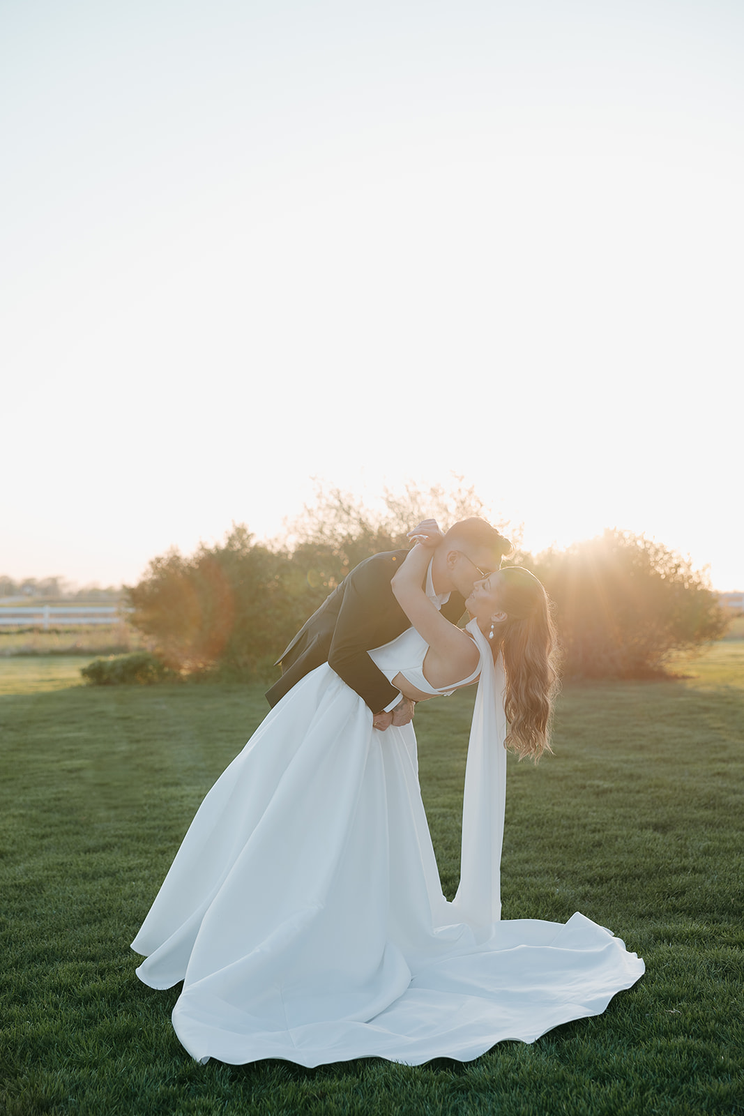 Groom dipping his bride for a kiss in pure golden hour light, her gown and cape pooling across the lawn in true old money wedding fashion
