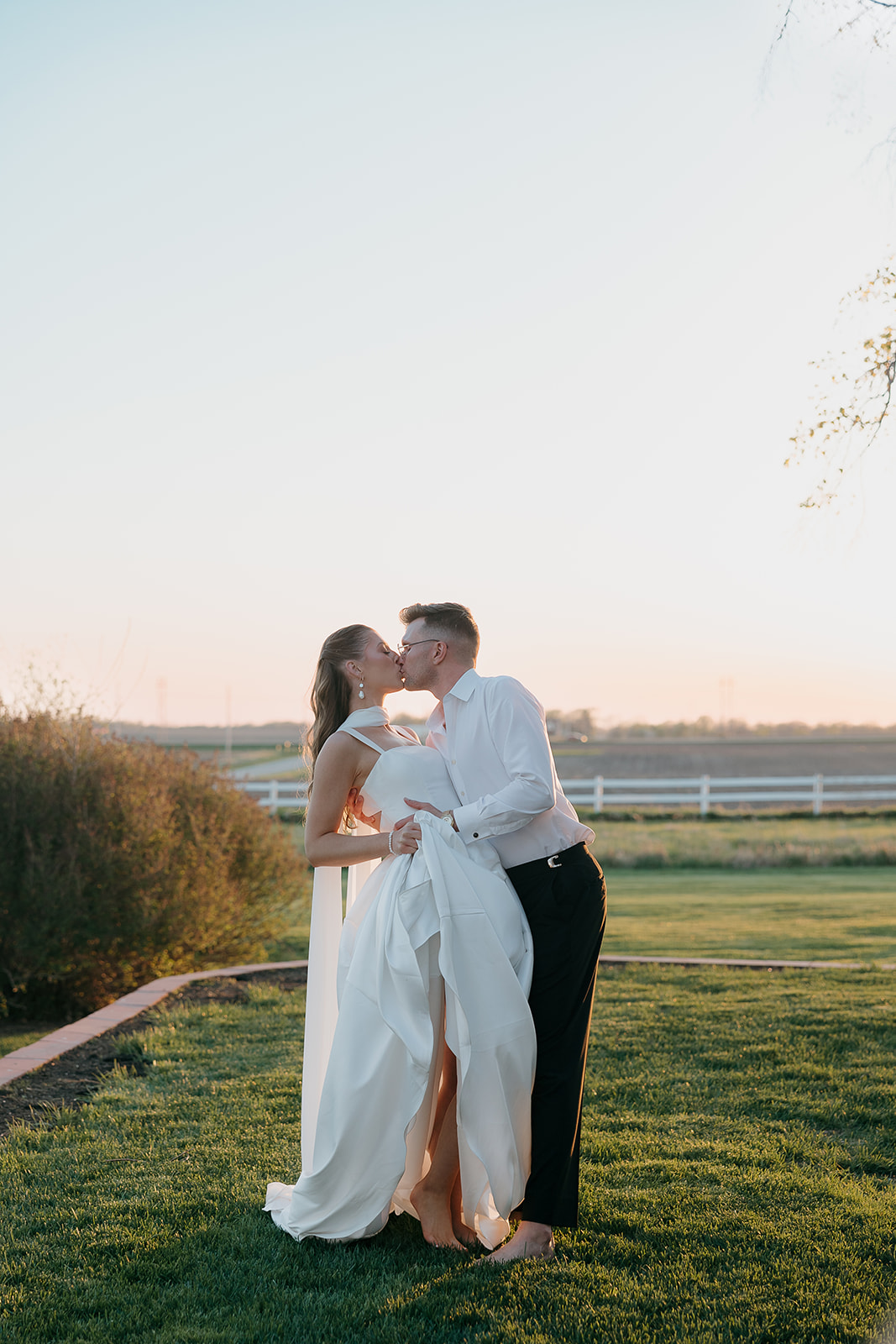 Barefoot bride and groom sharing a sunset kiss along a brick garden path, white fencing and open fields stretching behind them — effortlessly old money wedding at its best

