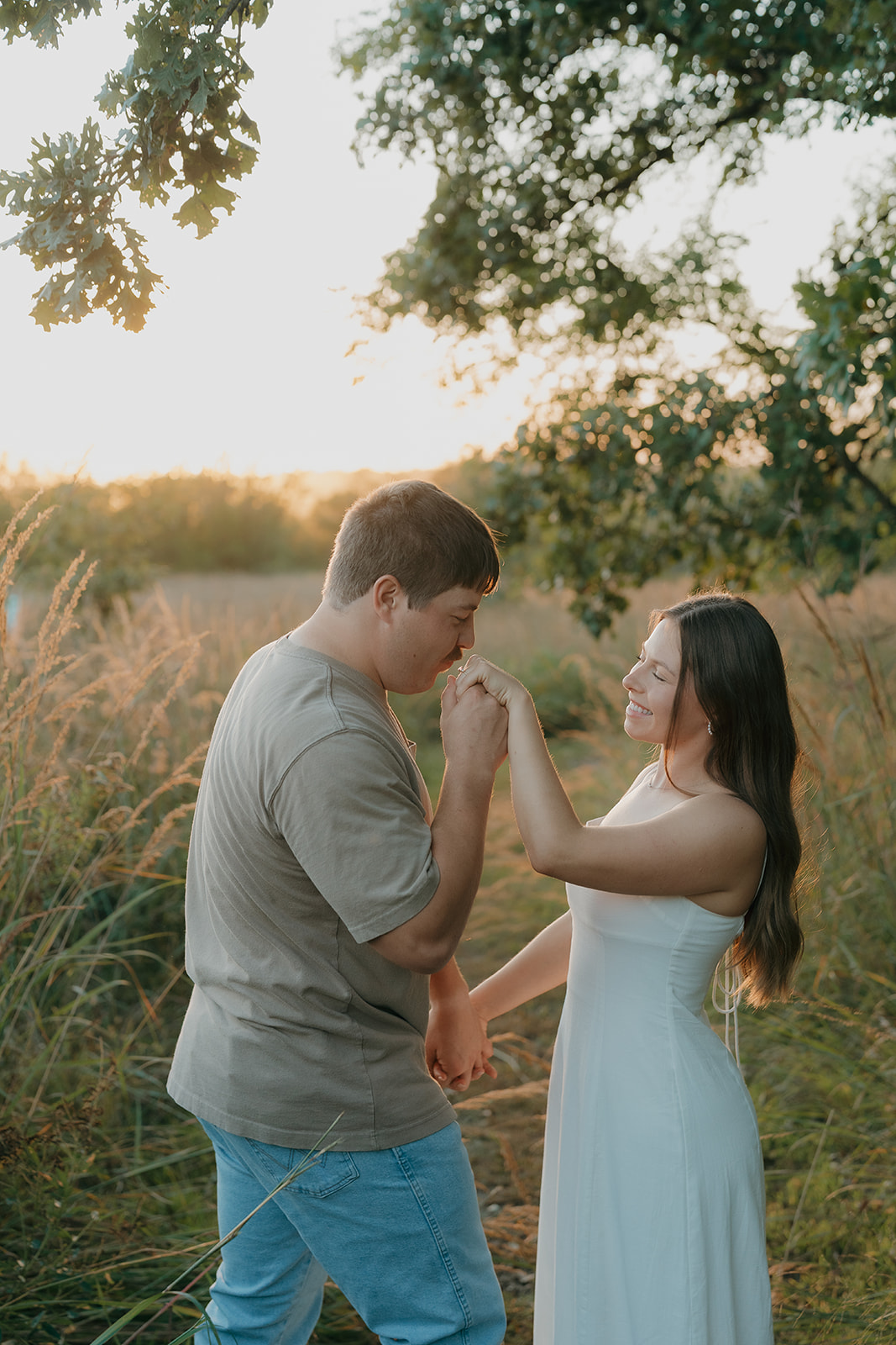 A man kisses his partner's hand while she laughs with eyes closed during an outdoor engagement session surrounded by tall golden grasses and glowing sunset light in Iowa