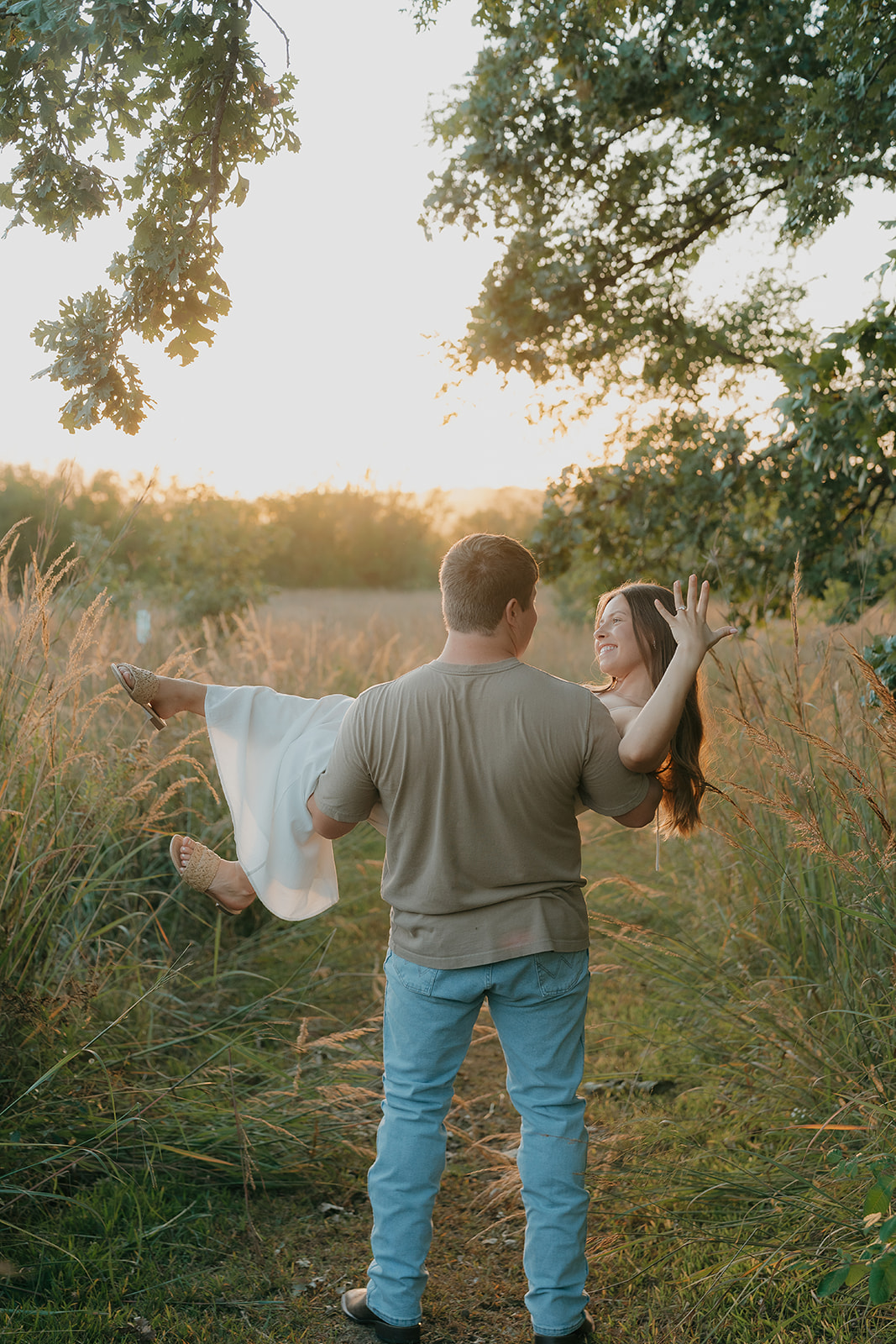 Couples photoshoot ideas with a man carrying his laughing partner through a glowing golden meadow at sunset with trees framing the scene