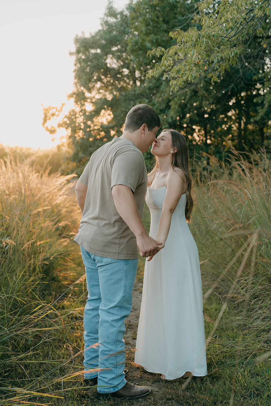 Couples photoshoot ideas in a tall grass prairie path as a man leans in close to his partner in a white dress while she smiles with eyes closed in warm golden hour light