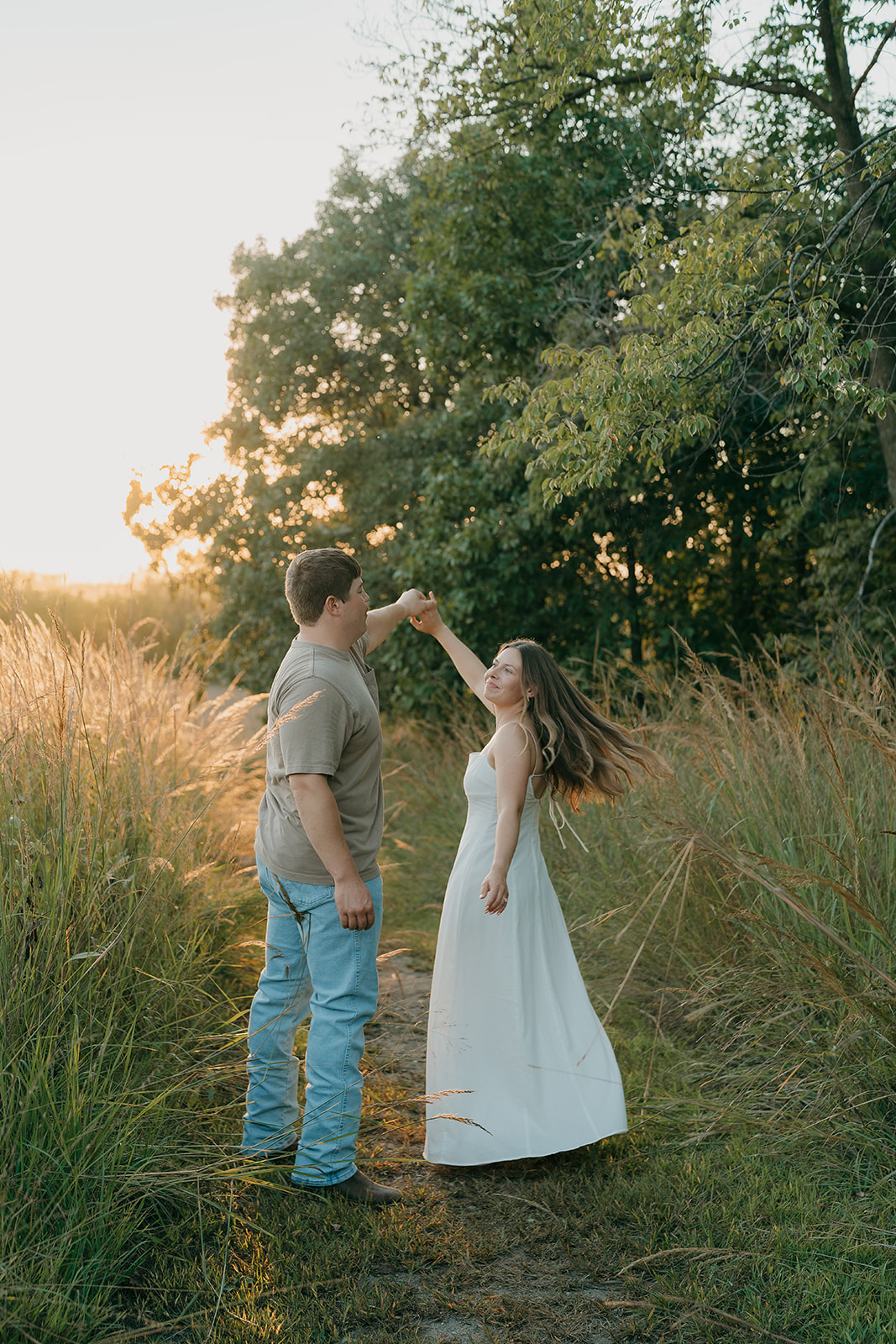 A man spins his partner in a white dress along a prairie path bordered by tall grasses with a vivid golden sunset and green trees glowing in the background