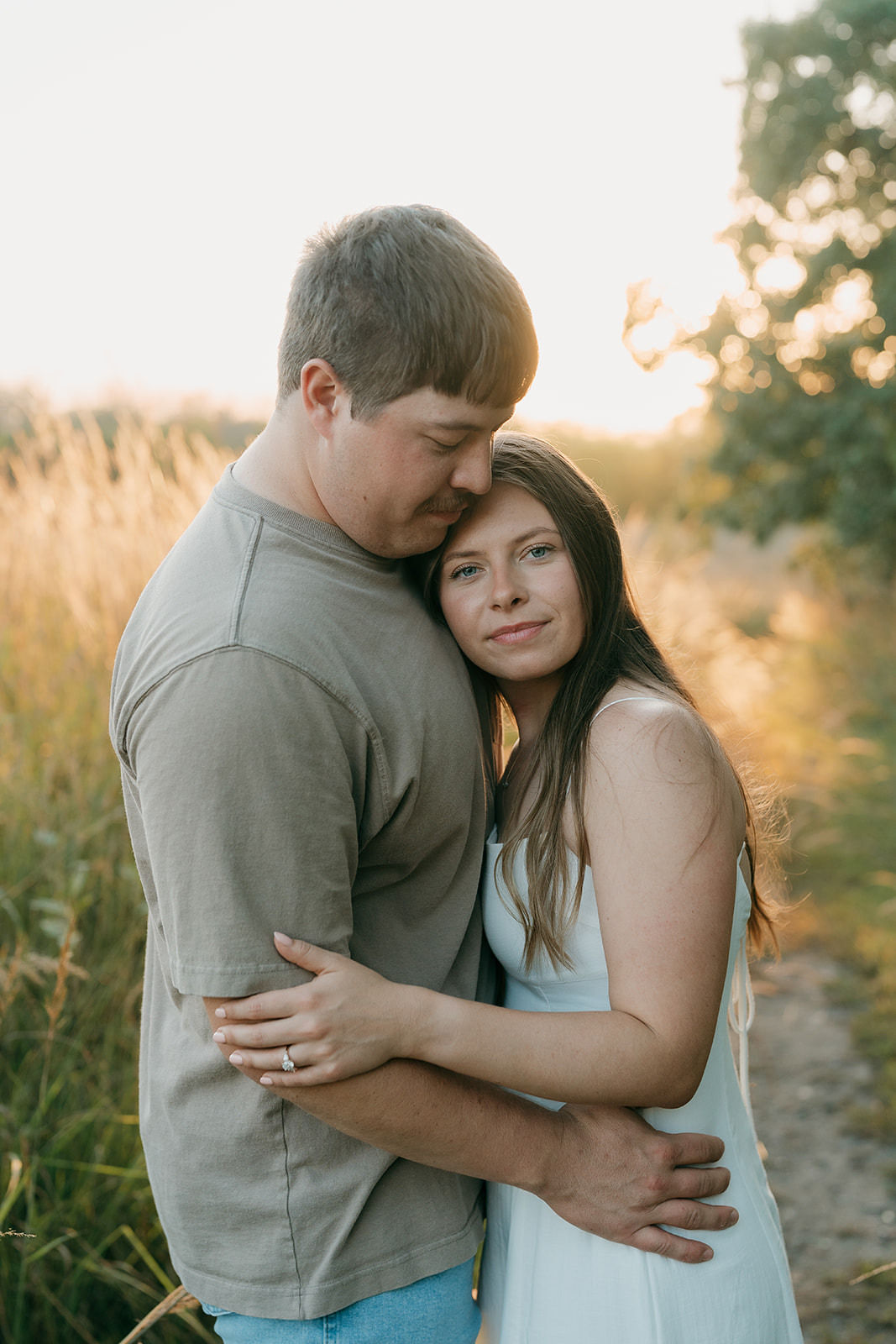 A woman in a white dress rests her head against her partner's chest as he holds her close in a sun-drenched field of tall grass at sunset