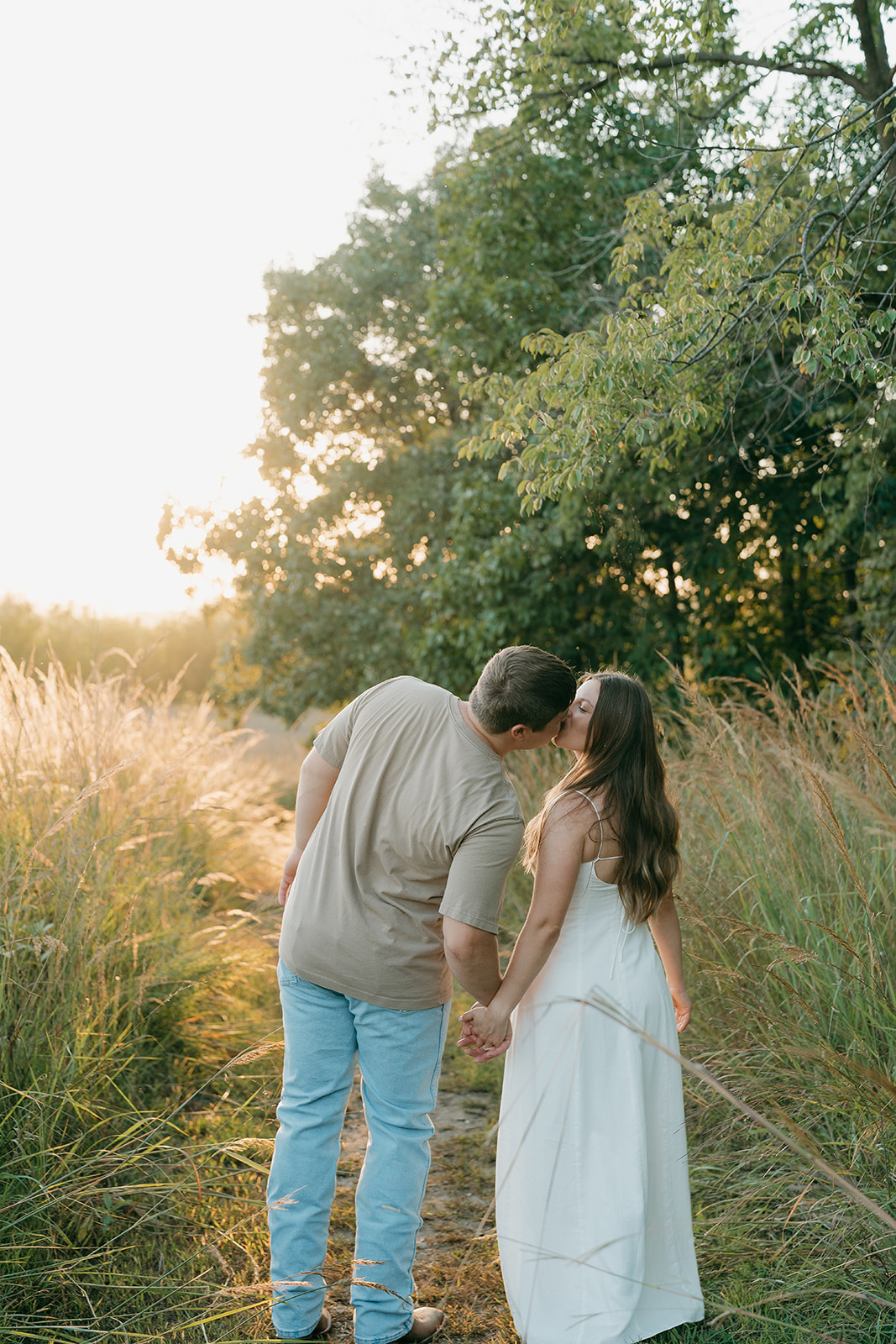 A couple shares a sweet kiss along a narrow prairie trail while holding hands with tall golden grasses on either side and soft warm light glowing through the trees ahead