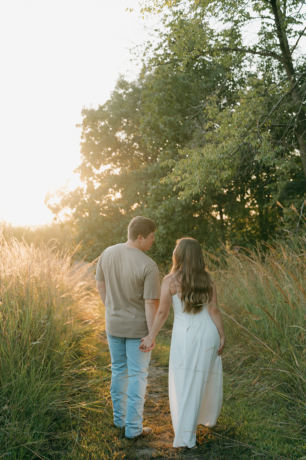 A couple holds hands walking away from the camera down a narrow trail bordered by tall grasses with warm backlit golden hour light filtering through the trees ahead