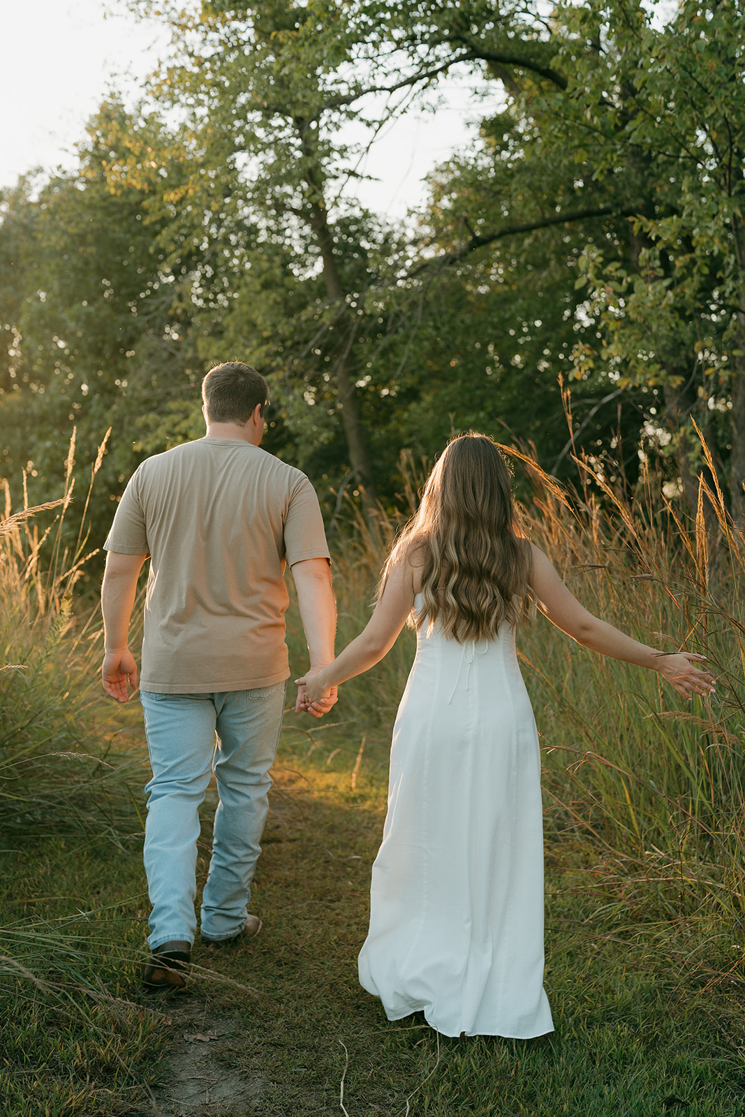 A couple walks hand in hand away from the camera through tall golden grasses at sunset with the woman in a white dress extending her arm freely at her side
