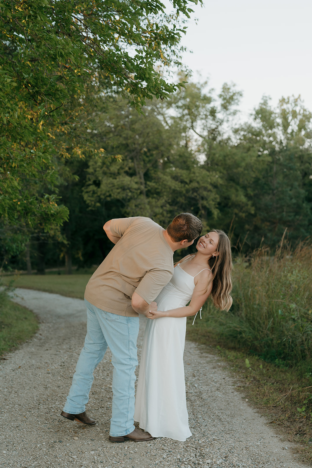Couples photoshoot ideas on a gravel path as a man leans in to kiss his laughing partner in a white maxi dress surrounded by lush green trees in Iowa
