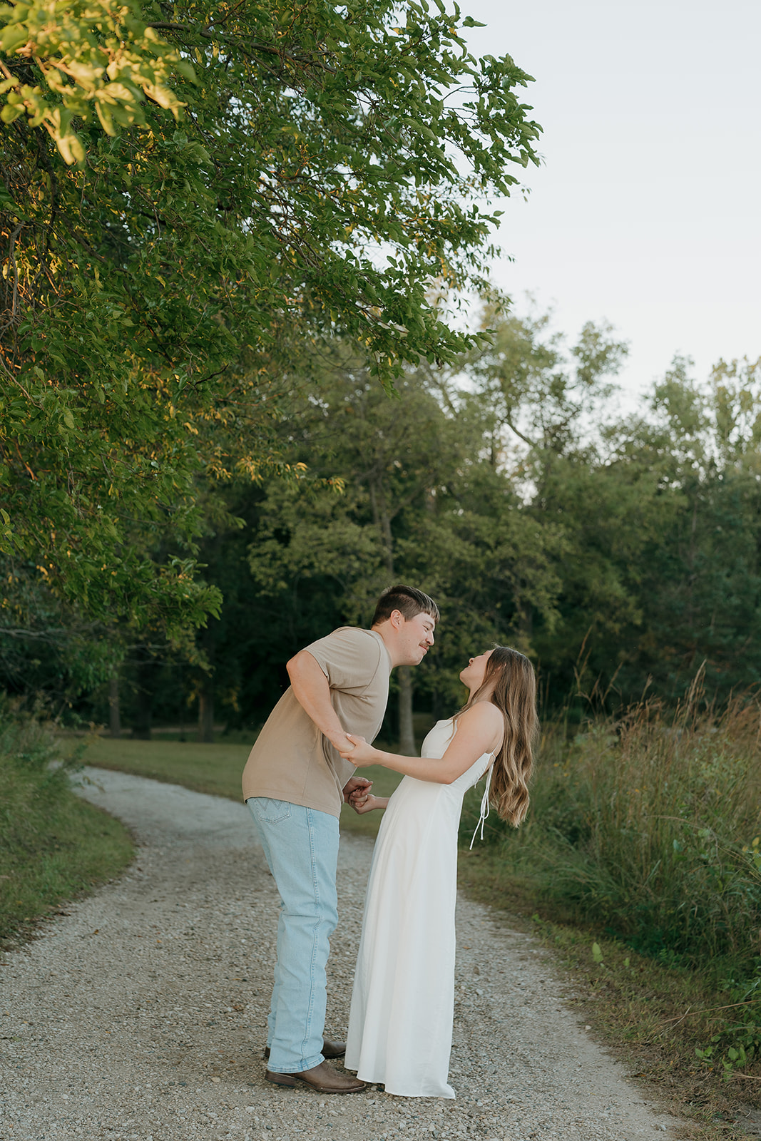 Couples photoshoot ideas on a gravel path as a man leans in smiling toward his partner in a white maxi dress with golden hour light filtering through the trees