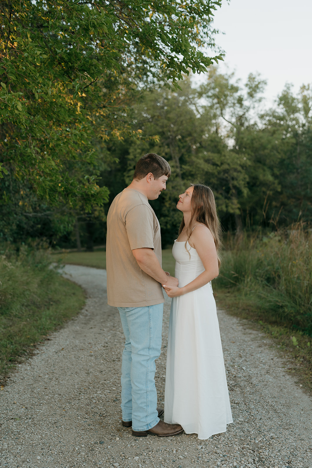 A man and woman stand facing each other on a gravel path holding hands and smiling softly with green trees and evening light surrounding them during an outdoor session in Iowa