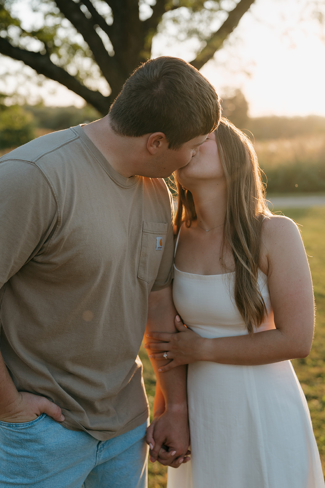 An engaged couple shares a tender kiss at golden hour beneath a large oak tree with warm sunset light glowing behind them and an engagement ring visible on the woman's hand