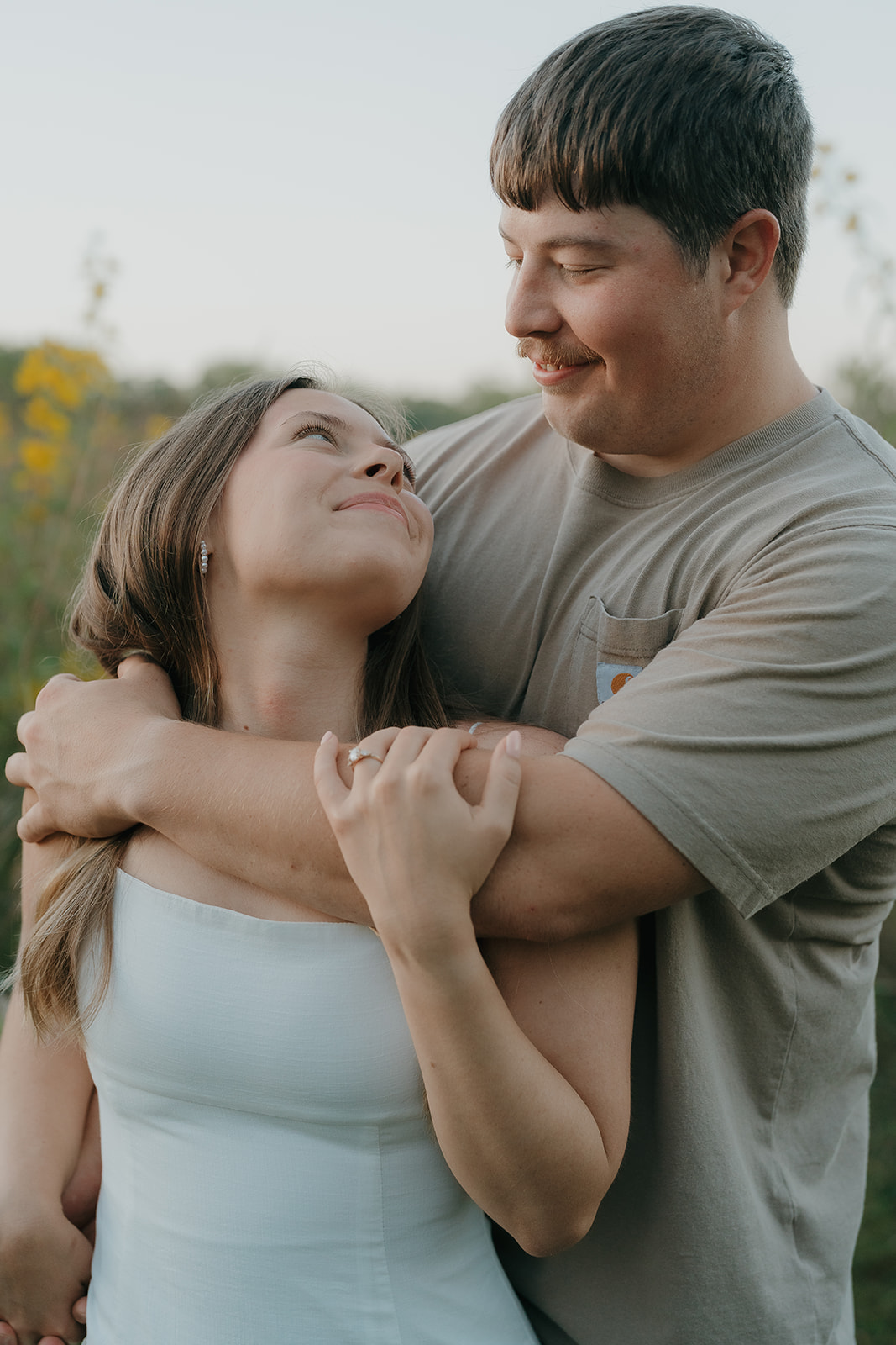 Couples photoshoot ideas featuring a close up of a man wrapping his arms around his partner from behind as she looks up at him smiling with her engagement ring visible and yellow wildflowers in the background