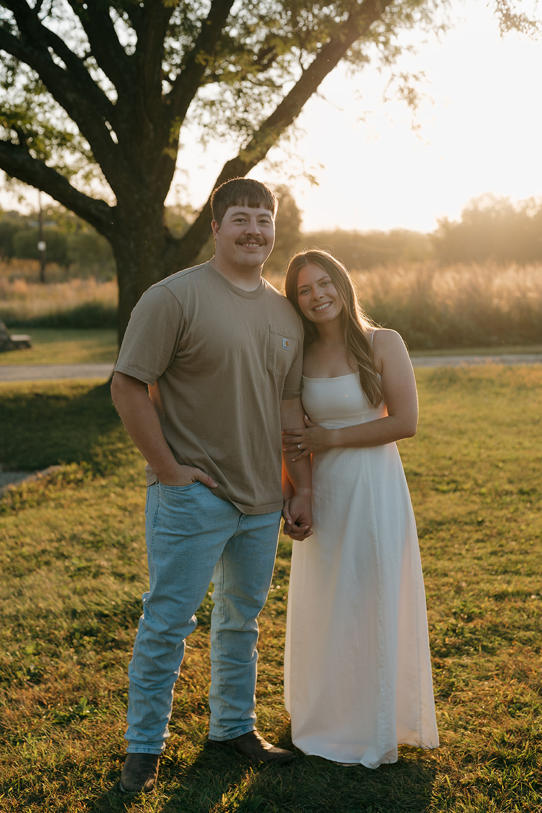 Couples photoshoot ideas with an engaged couple smiling at the camera while standing side by side beneath a large oak tree with warm golden sunset light glowing behind them in Iowa