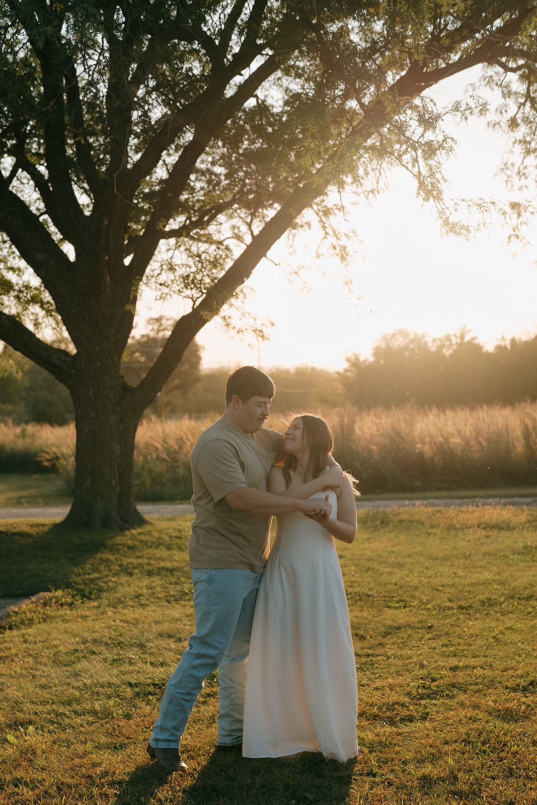 A couple dances and laughs together beneath a large oak tree with warm golden sunset light glowing behind them in an open Iowa field