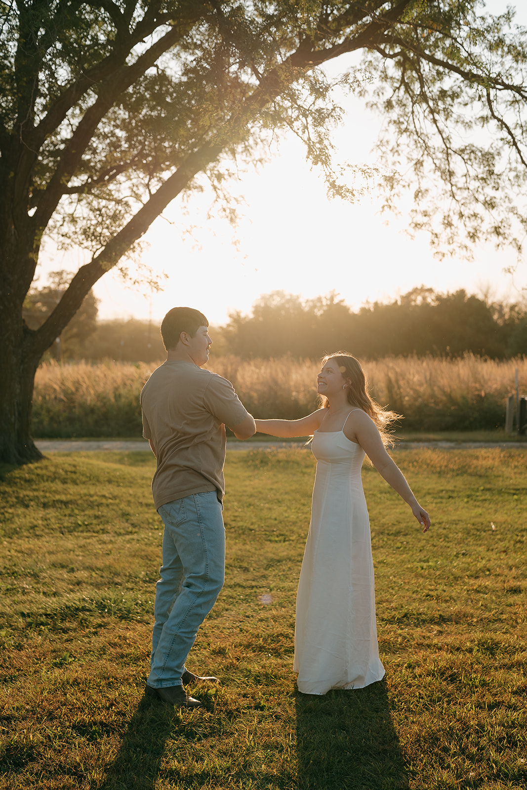 ouples photoshoot ideas showing a man and woman dancing together in an open field with a large tree silhouetted against a blazing golden sunset and tall prairie grasses in the background