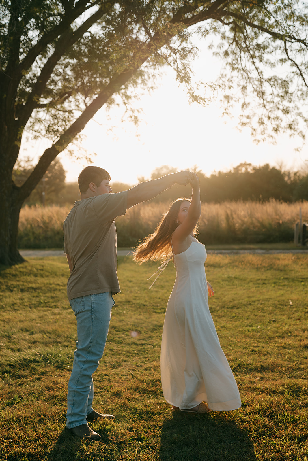Couples photoshoot ideas showing a man spinning his partner in a white dress under his raised arm in an open field with a glowing golden sunset and large oak tree behind them