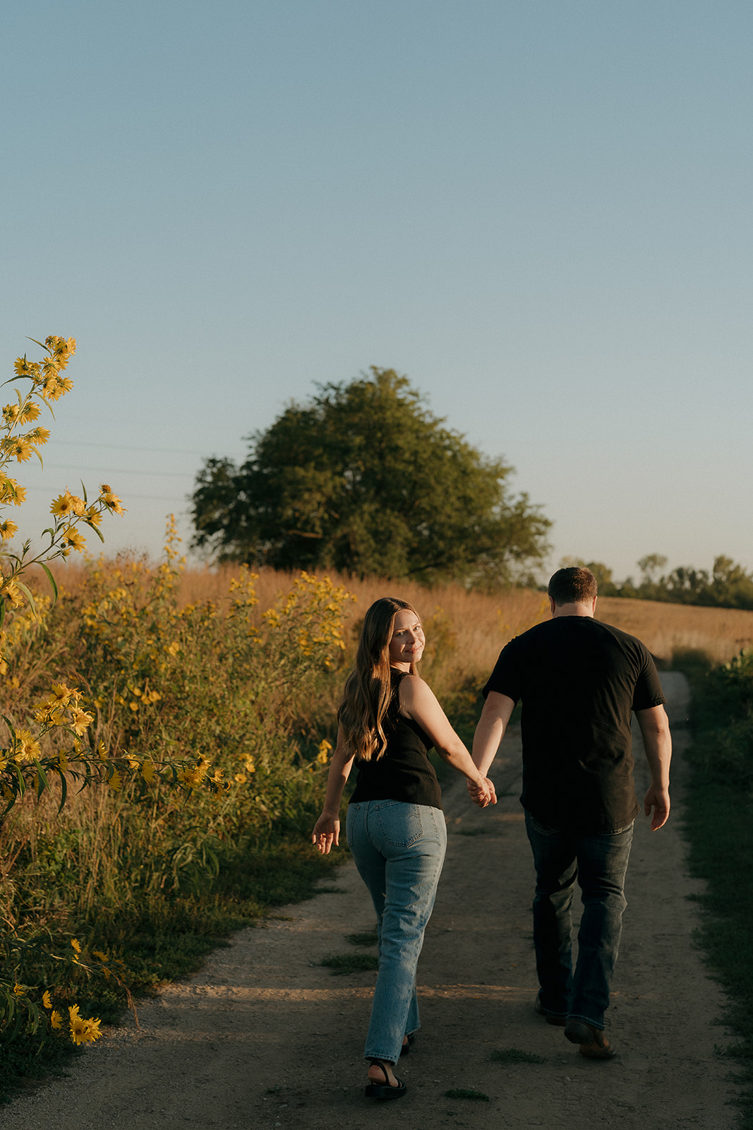 Couples photoshoot ideas along a country path lined with yellow wildflowers as the woman glances back over her shoulder with a smile while holding her partner's hand at sunset