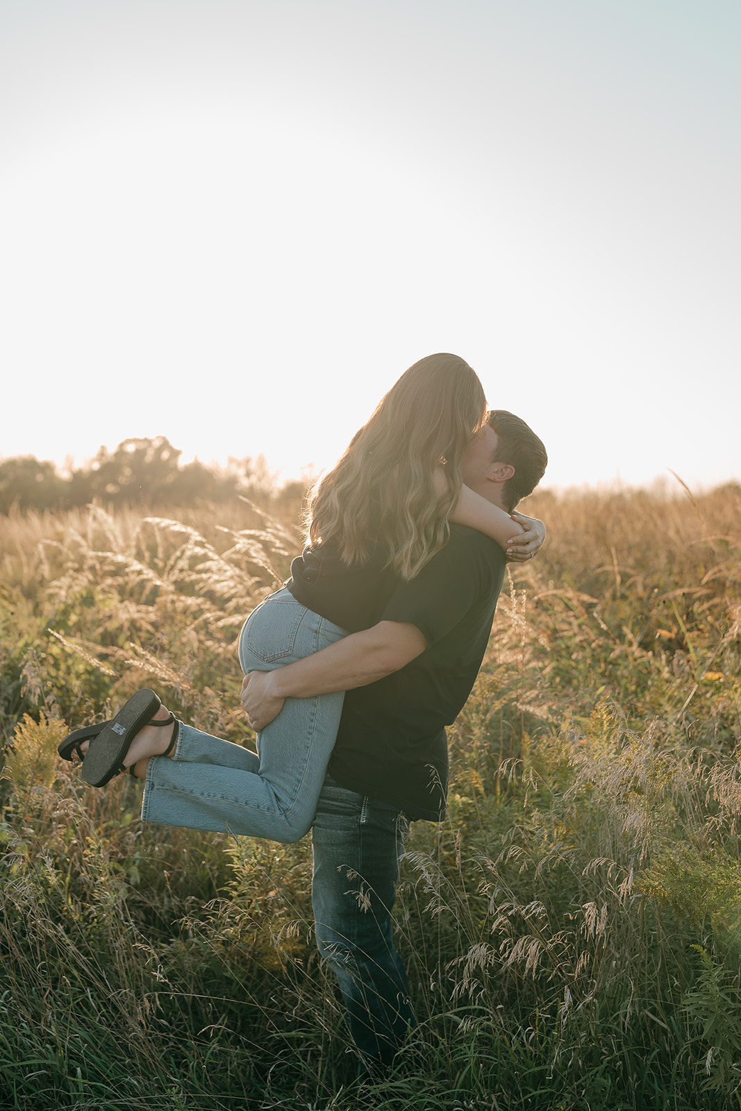 Couples photoshoot ideas in a tall grass prairie field as a man lifts his partner into a kiss with bright backlit golden hour light surrounding them
