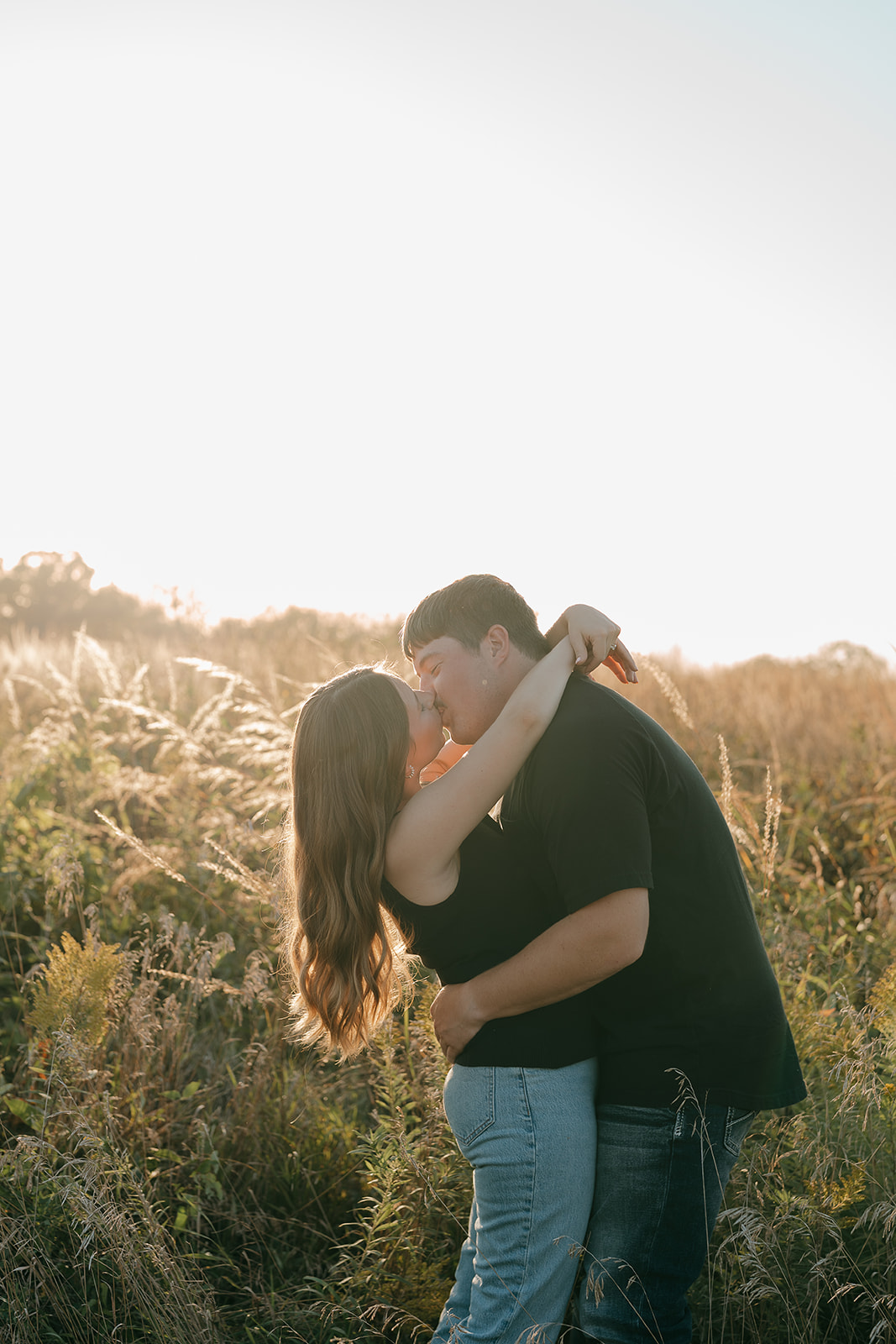 A couple kisses in a sunlit field of tall prairie grasses with bright golden light bursting behind them and the woman's arm wrapped around the man's neck