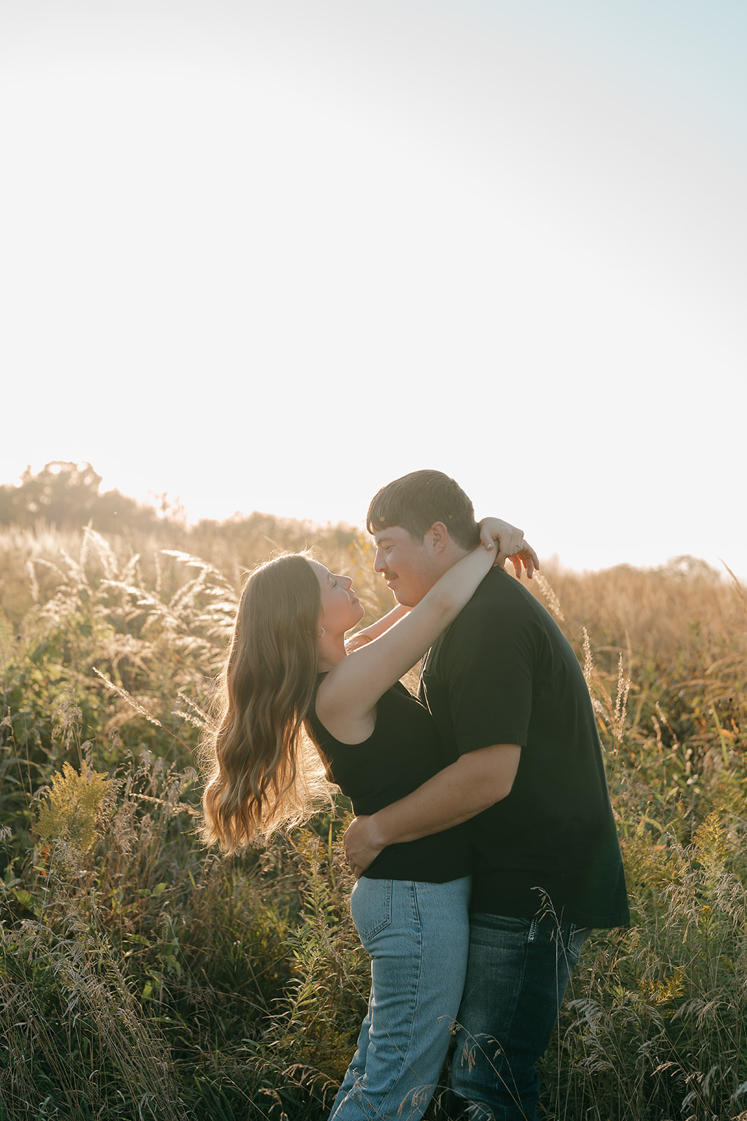 A couple embraces in a sunlit prairie field with the woman wrapping her arm around the man's neck as they look at each other with bright backlit golden light behind them
