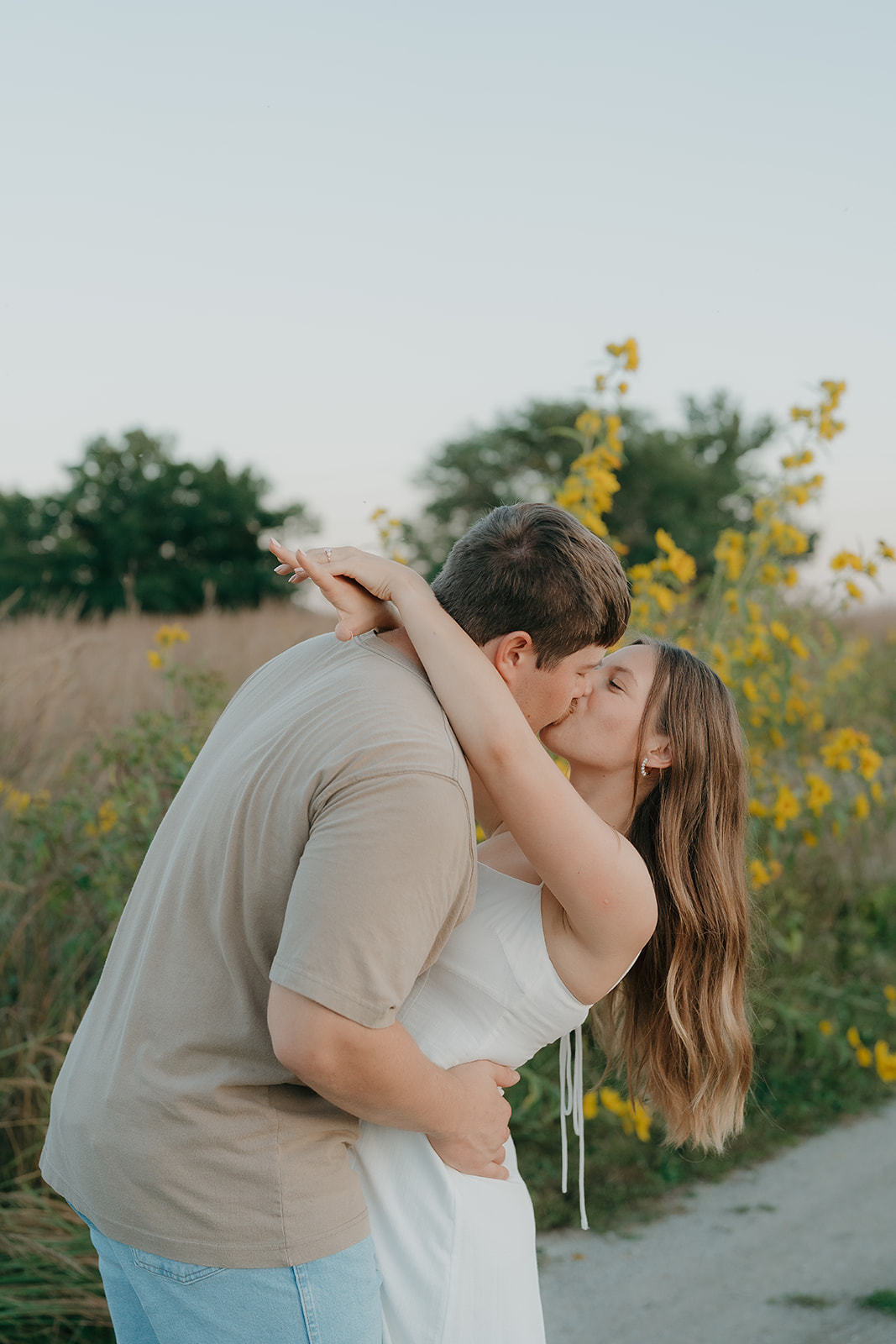 An engaged couple shares a kiss surrounded by tall golden grasses and yellow wildflowers during a summer evening outdoor session in Iowa