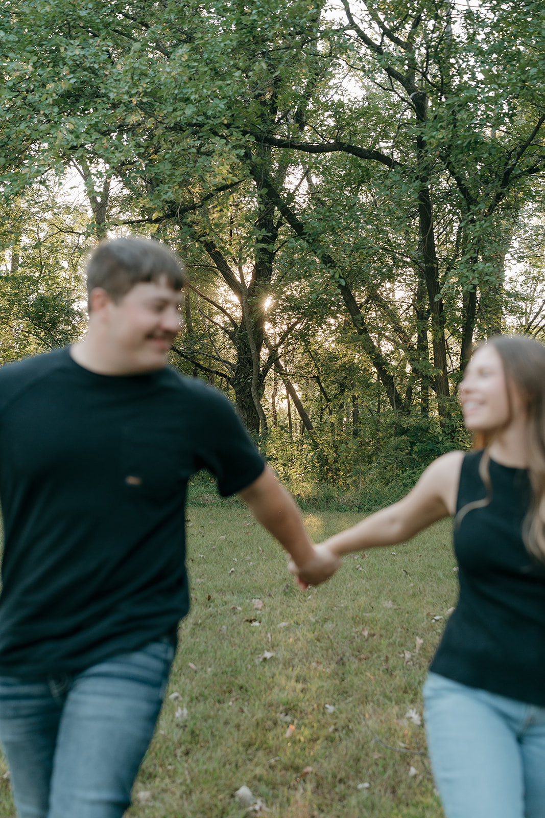 A smiling couple holds hands and walks through a grassy wooded clearing while laughing and looking at each other with golden sunset light breaking through the trees behind them