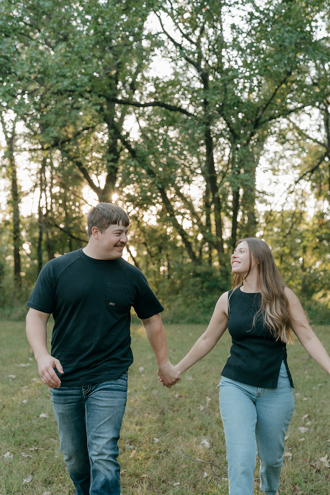 Couples photoshoot ideas featuring a laughing man and woman holding hands and walking through a sunlit wooded clearing while smiling at each other with sunset light glowing through the trees