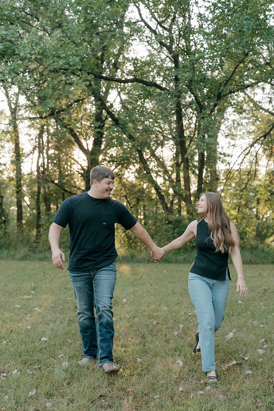 Couples photoshoot ideas with a laughing couple in matching black outfits holding hands and walking through a grassy field while smiling at each other with evening light filtering through the trees