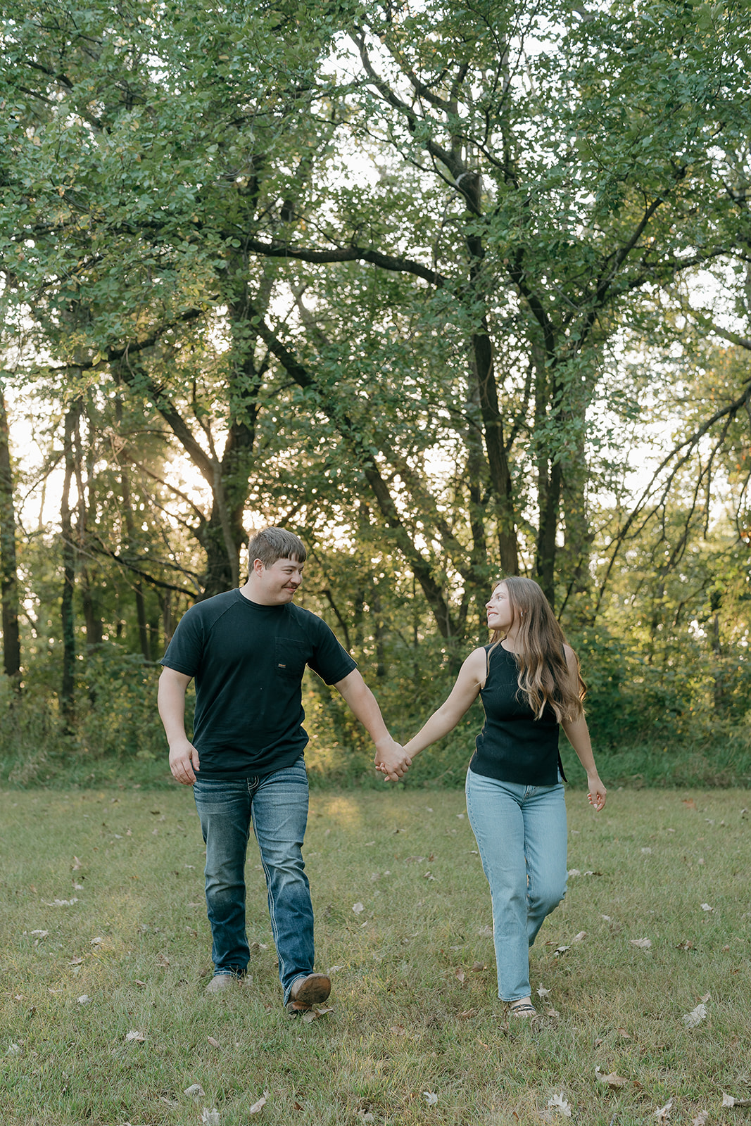 Couples photoshoot ideas featuring a man and woman holding hands and walking through a sunlit park while smiling at each other with lush green trees in the background