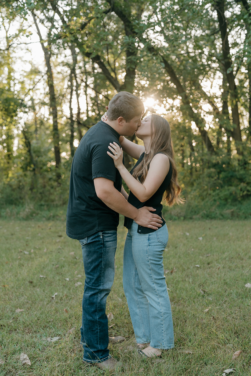 A couple shares a kiss in a wooded clearing with soft sunlight breaking through the trees behind them during an outdoor engagement session in Iowa