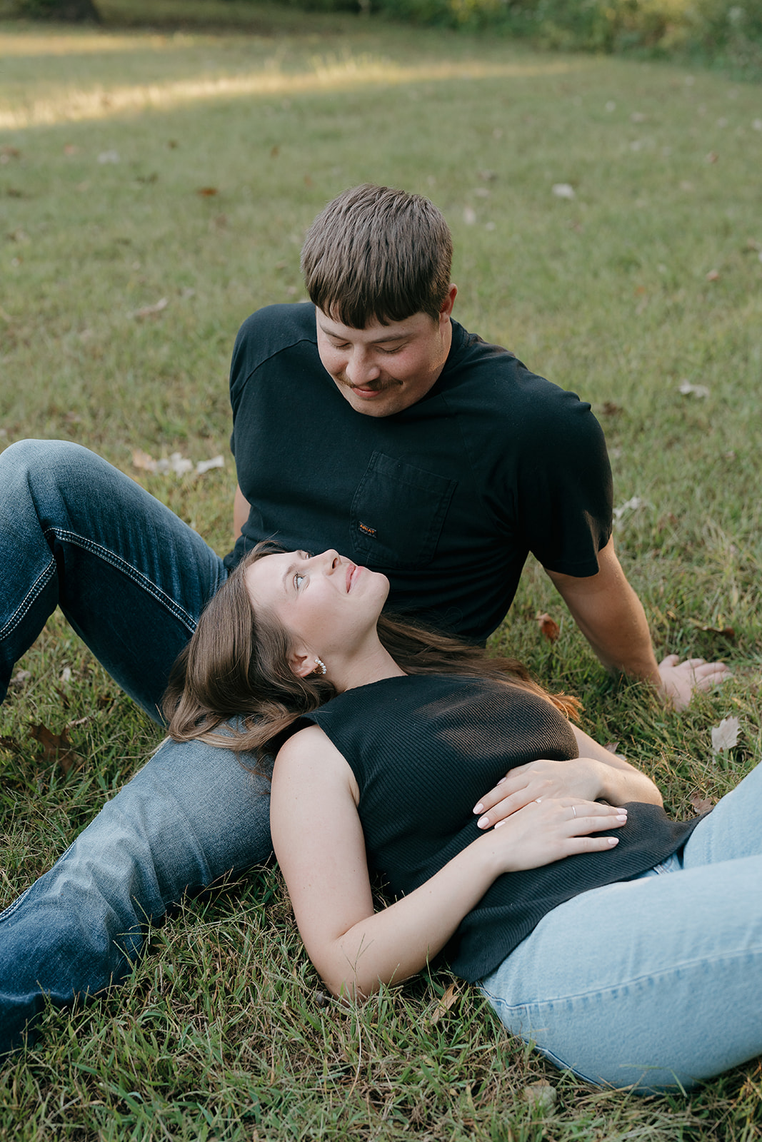 A woman lies in the grass looking up at her partner who sits beside her smiling down at her during an outdoor engagement session in Iowa