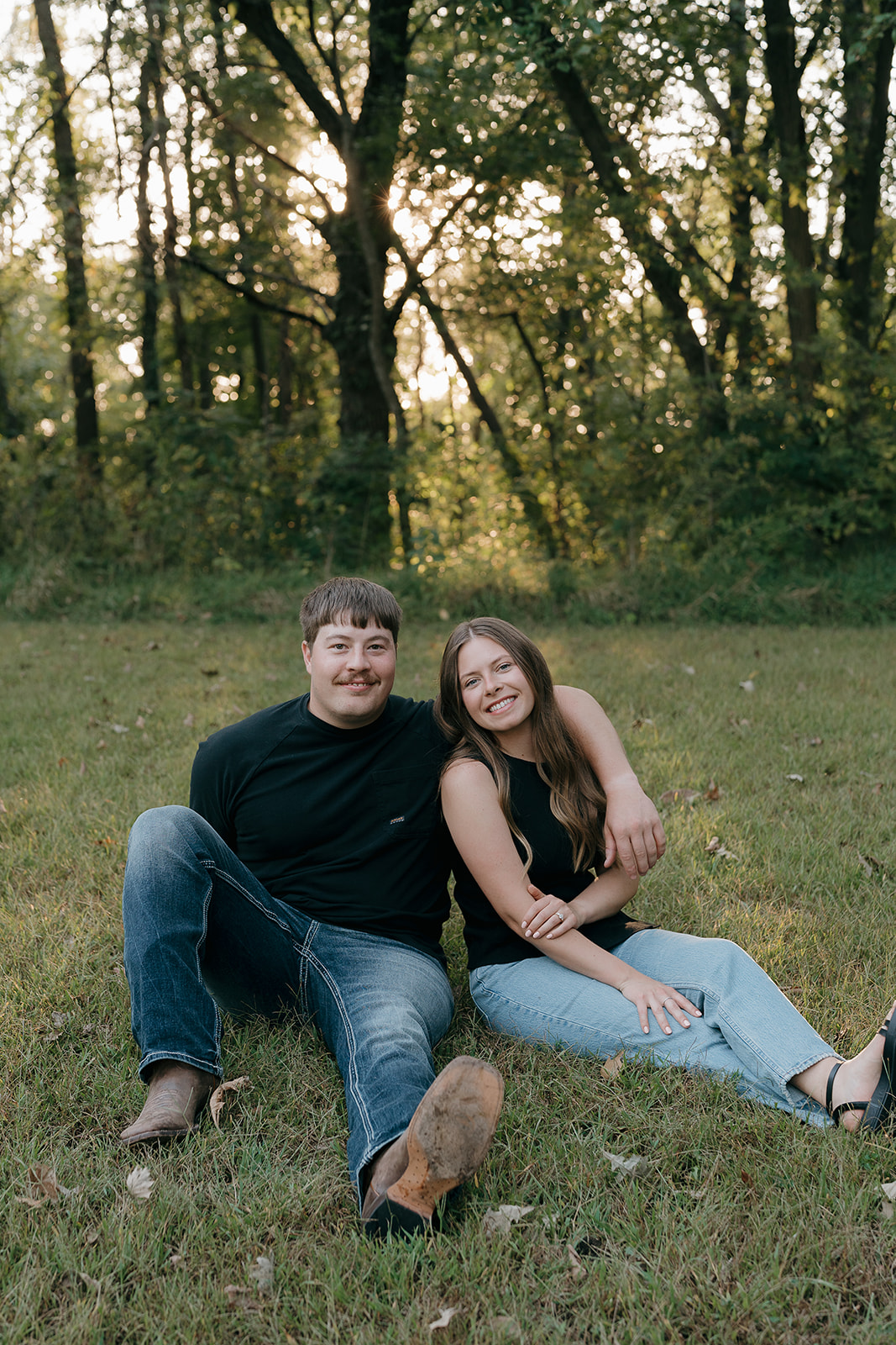 A smiling couple sits together in the grass in matching black outfits looking at the camera with golden light breaking through the trees behind them during an Iowa engagement session