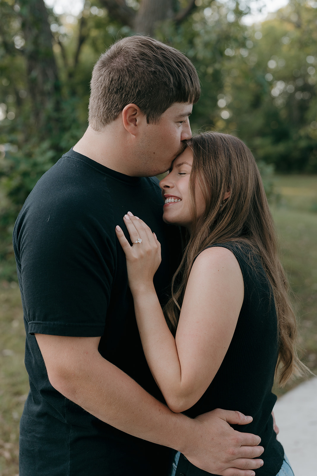 Couples photoshoot ideas with a man kissing his partner on the forehead as she smiles and presses her hand with her engagement ring against his chest in a wooded park setting