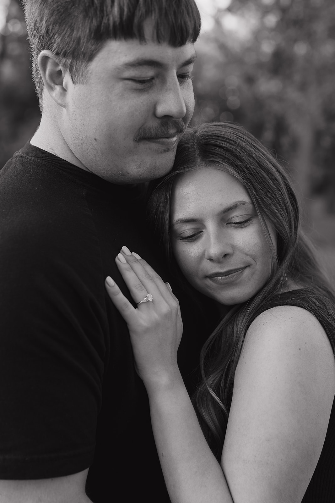 Couples photoshoot ideas in black and white showing a woman leaning against her partner's chest with eyes closed and her engagement ring visible on his chest