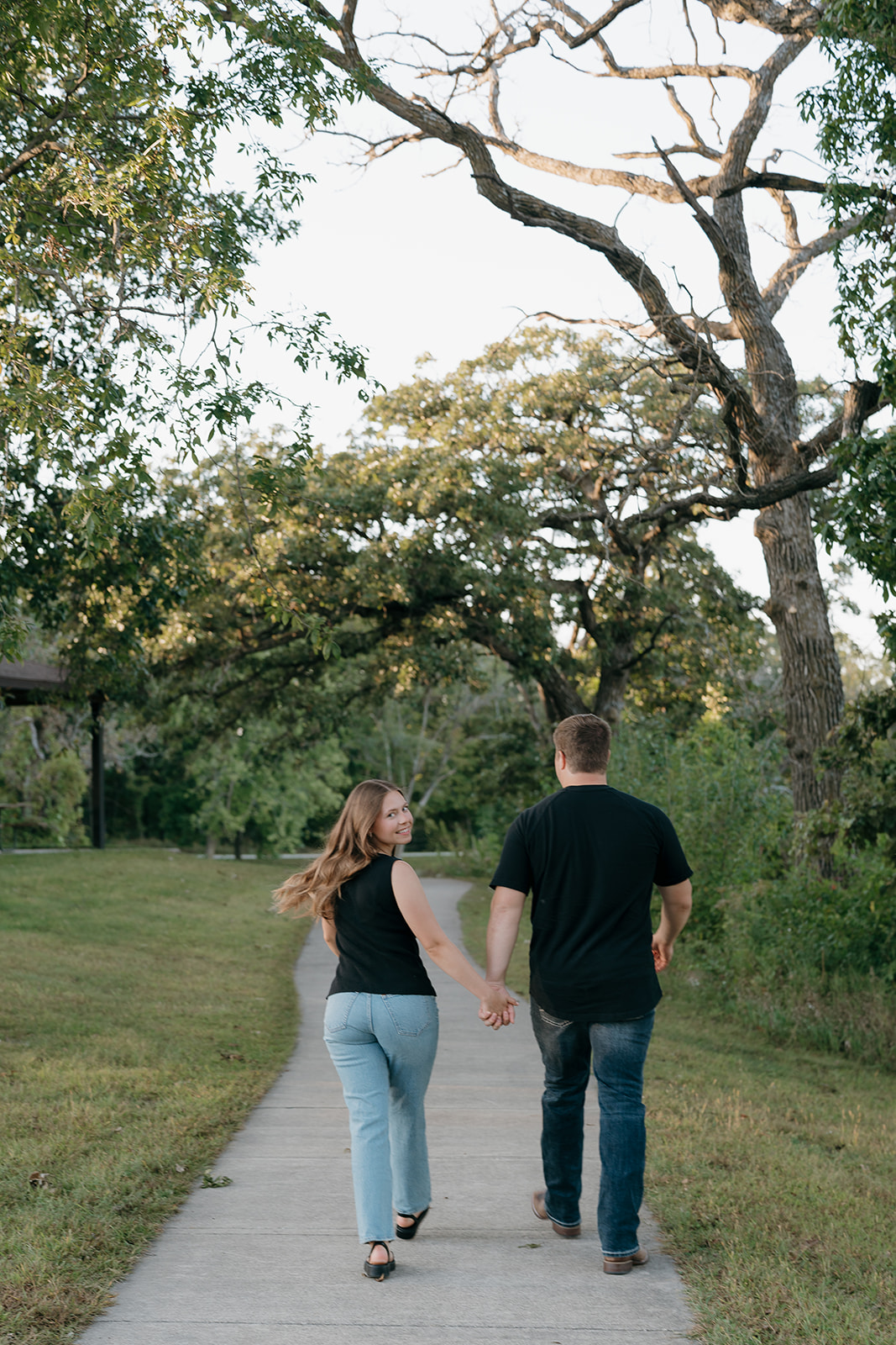 A couple walks hand in hand down a tree-lined path away from the camera as the woman glances back over her shoulder with a smile
