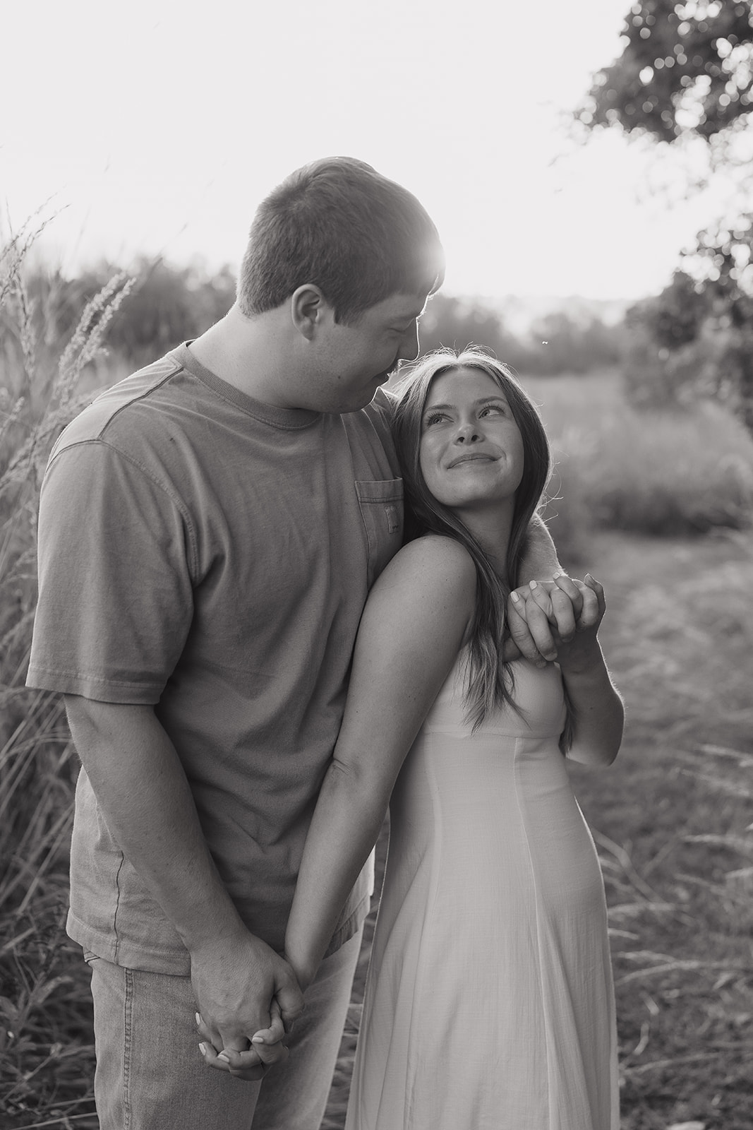 Couples photoshoot ideas in black and white showing a woman leaning against her partner's arm and smiling up at him while he looks down at her tenderly in a field of tall grass