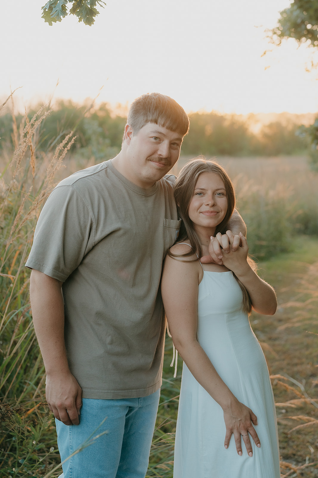 An engaged couple stands together in a sun-drenched prairie field smiling at the camera with the woman resting her head on the man's shoulder and golden light wrapping around them