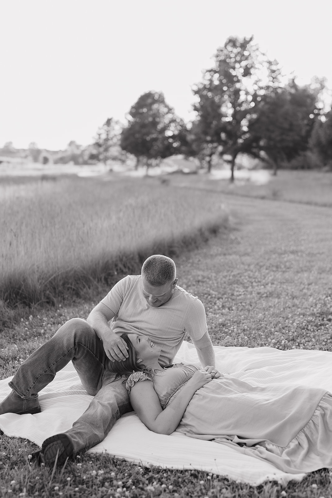 A black and white photo of the couple relaxing on a blanket in a meadow, the man leaning tenderly over his fiancée as she reclines in the grass

