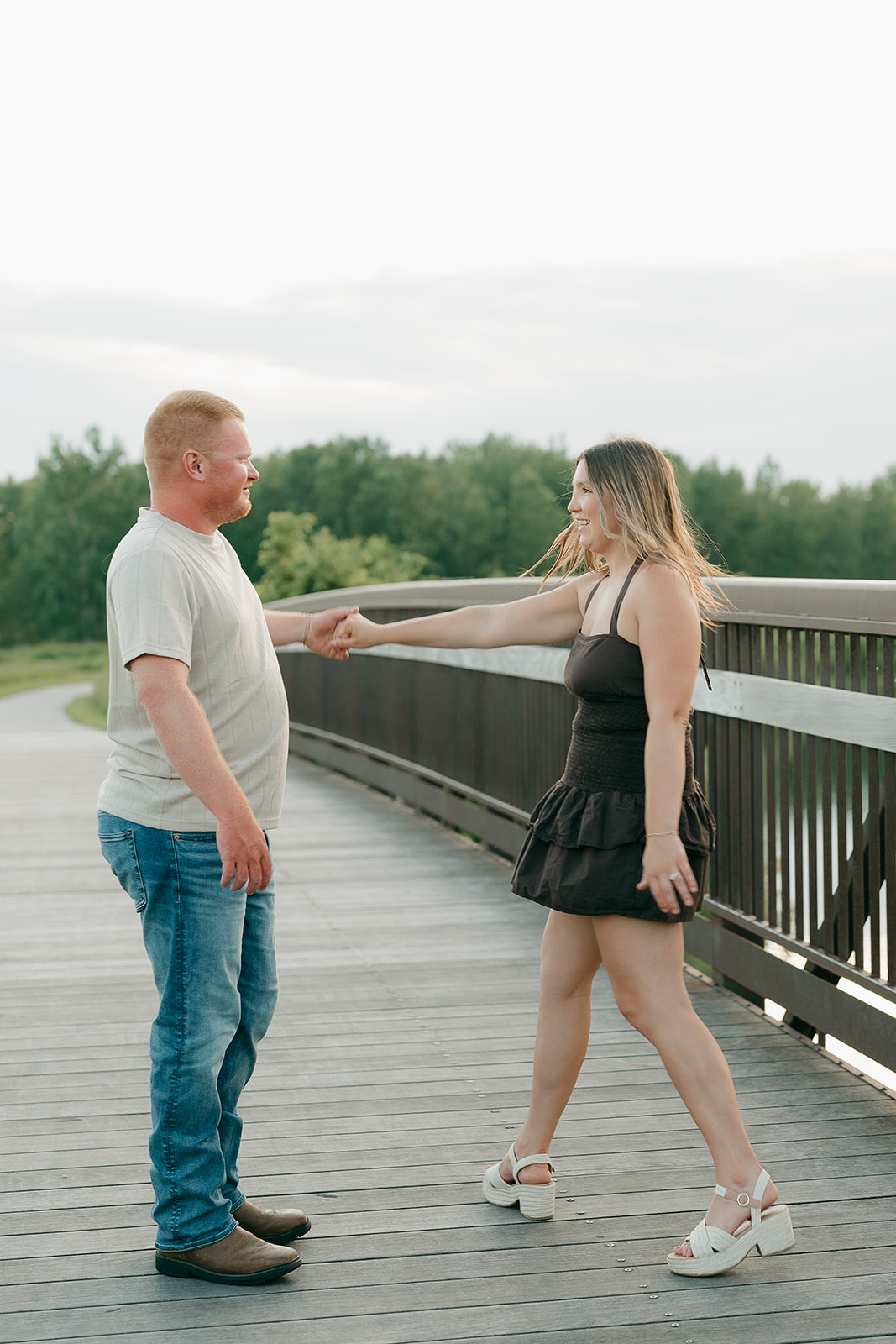The woman takes her partner's hand on the curved wooden bridge at Ada Hayden, her dark smocked dress and white platform sandals catching the evening light
