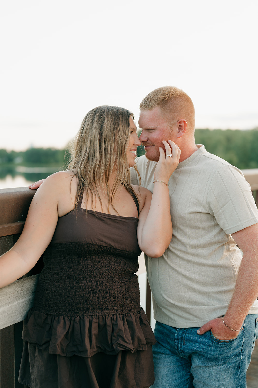 The couple leans close on a bridge railing at Ada Hayden, the woman in a dark smocked dress cradling her partner's face to show off her engagement ring
