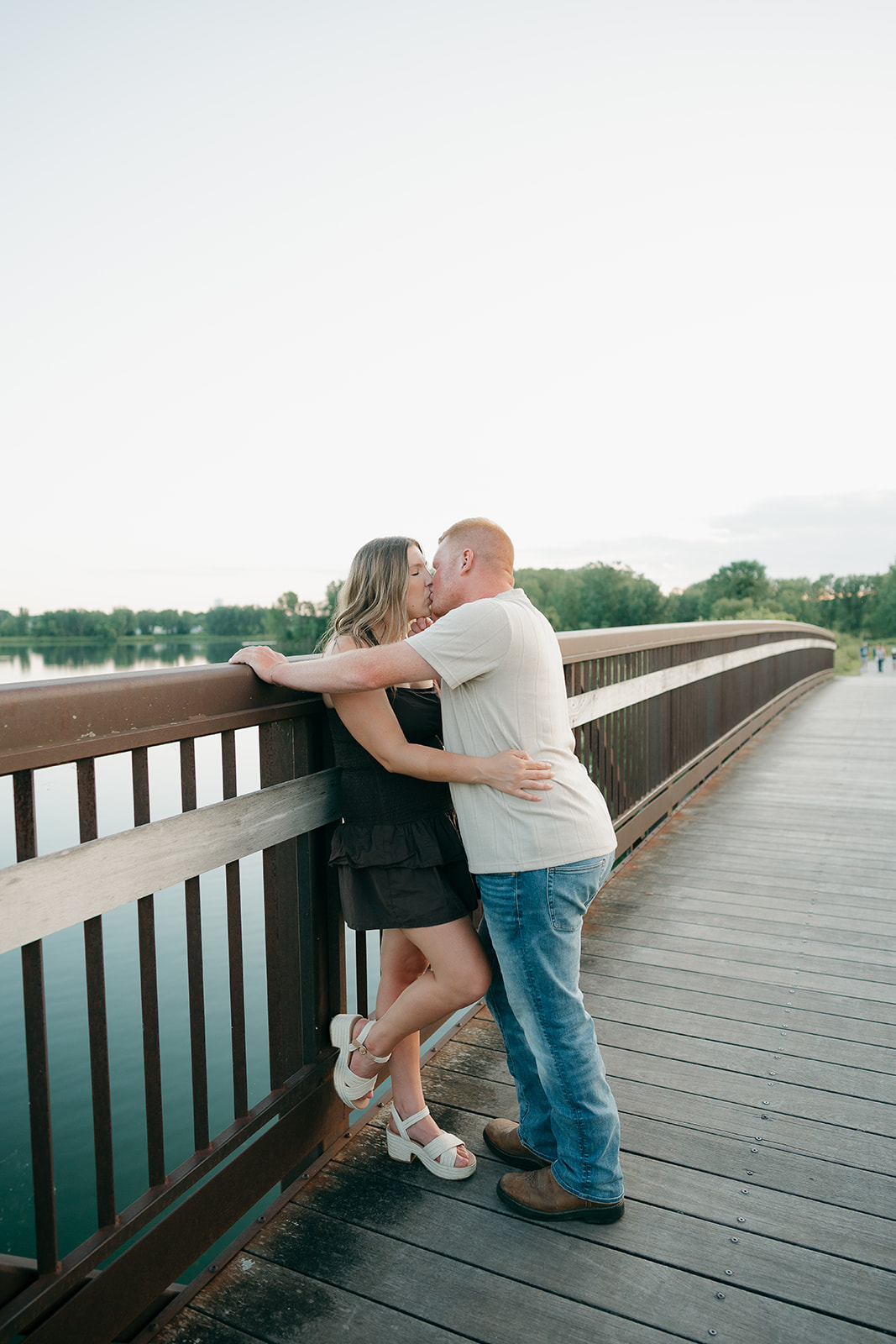 A couple shares a romantic kiss leaning against the railing of the wooden bridge at Ada Hayden, her dark dress and white sandals a classic summer engagement photo outfit pairing
