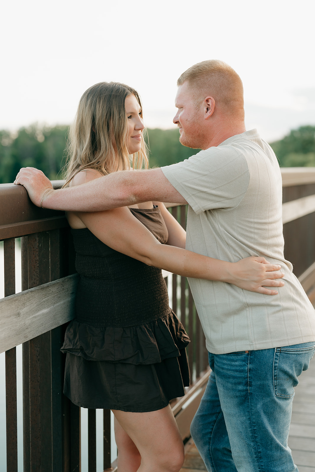 A couple wearing summer engagement photo outfits faces each other on a wooden bridge at Ada Hayden, smiling and holding each other close in the warm evening light
