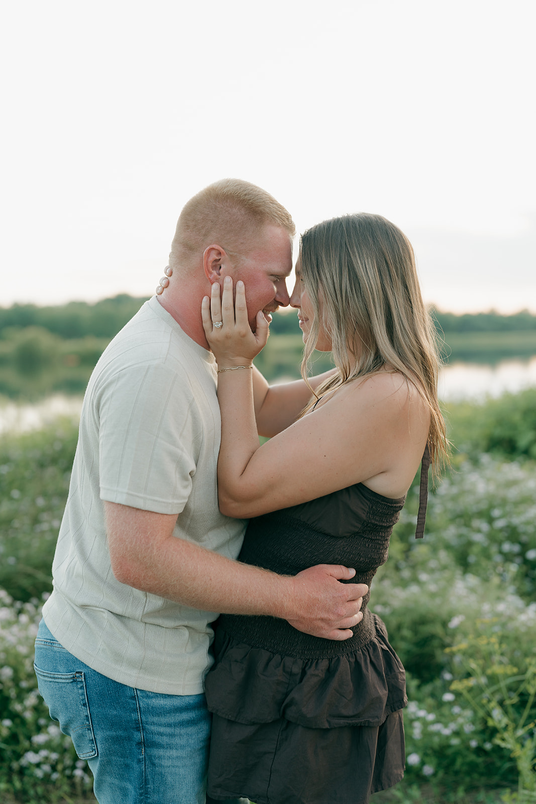 The couple leans forehead to forehead in a wildflower meadow beside Ada Hayden lake, the woman's engagement ring visible as she holds her partner's face
