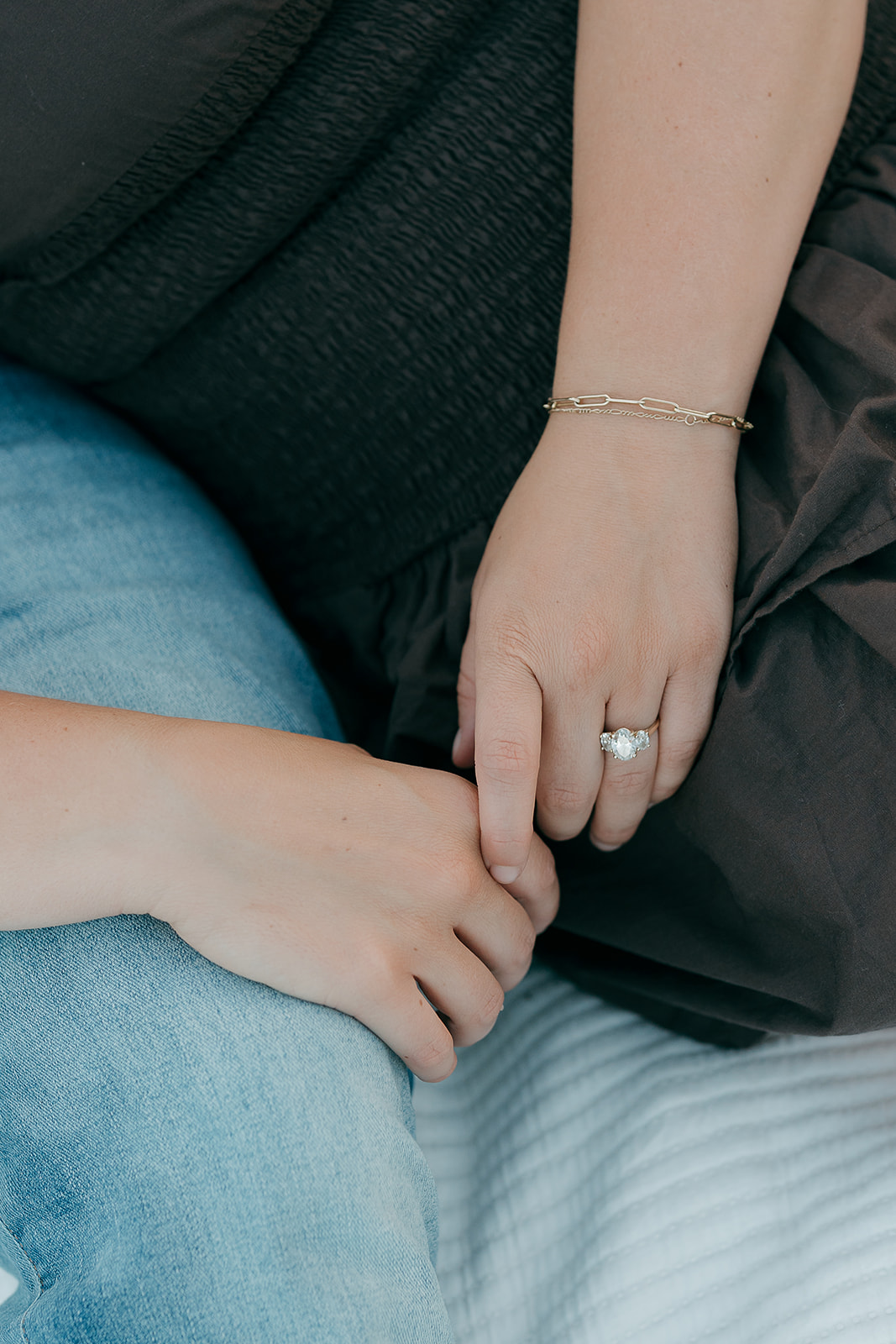 A close-up detail shot of the woman's oval diamond engagement ring and gold chain bracelet resting against her dark smocked dress during the Ada Hayden engagement session
