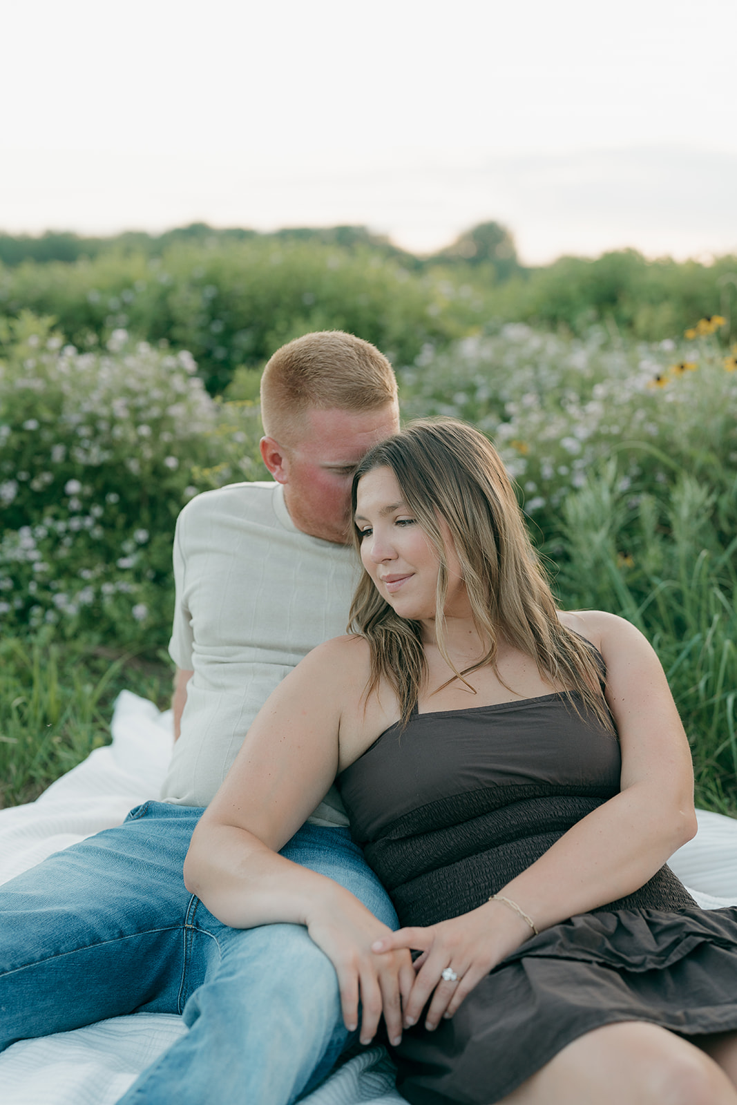 A couple relaxes on a blanket in a field of purple wildflowers and black-eyed Susans, the woman in a dark summer dress leaning into her partner's shoulder
