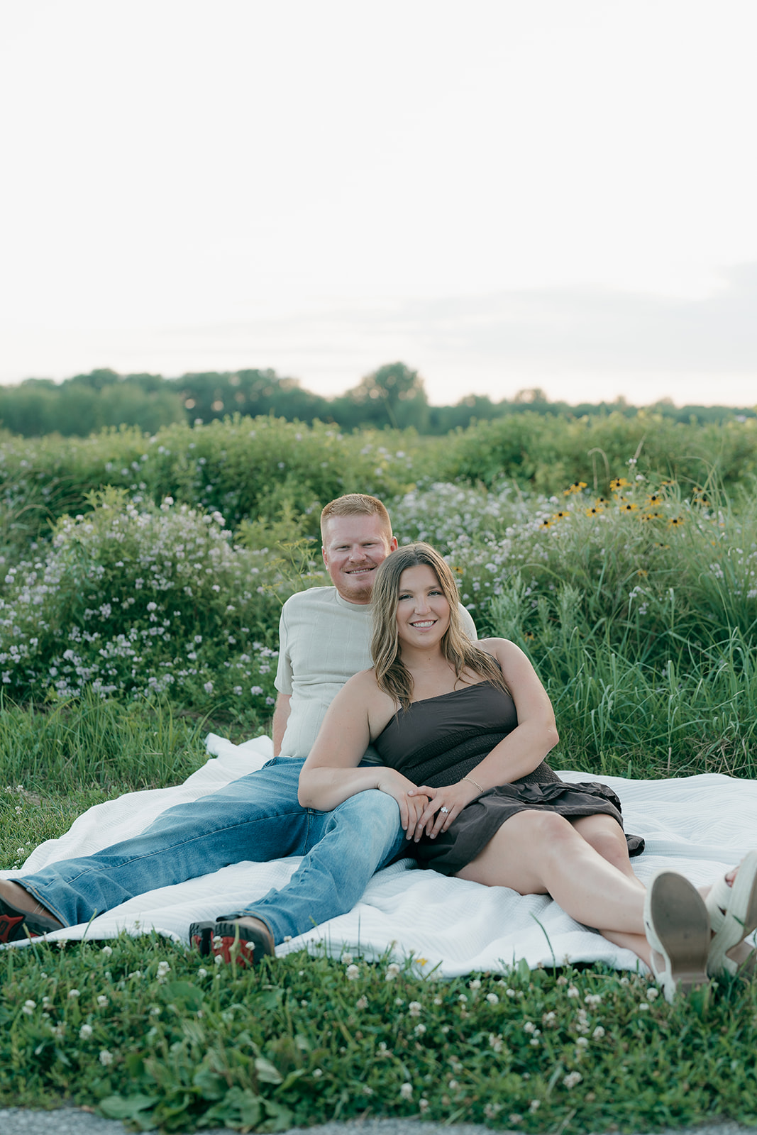 A couple in summer engagement photo outfits smiles at the camera while sitting together on a blanket in front of a wildflower field full of purple blooms and black-eyed Susans
