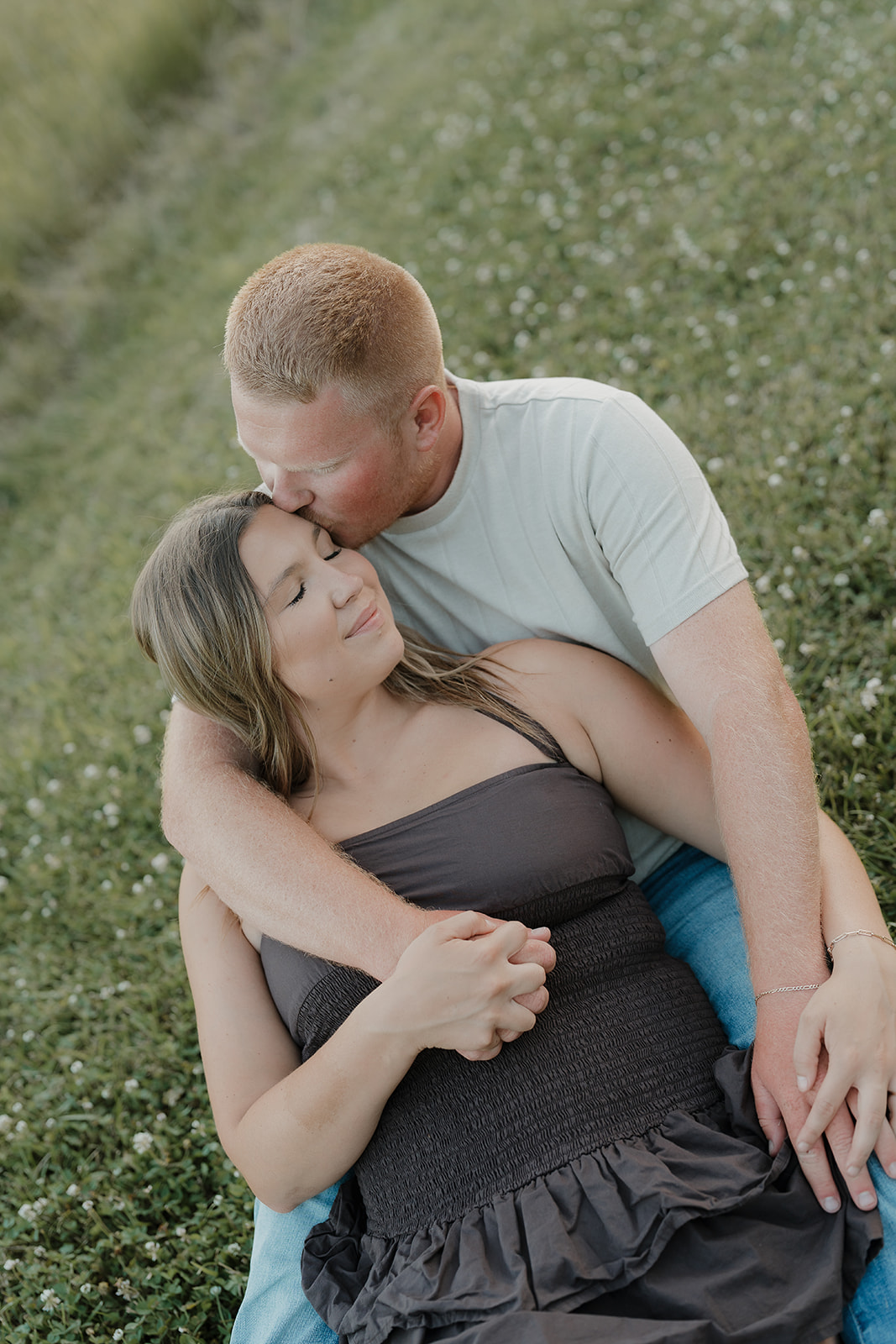 The couple sits close together in a wildflower field, the woman in a dark smocked dress leaning back against her partner as he gently kisses her forehead
