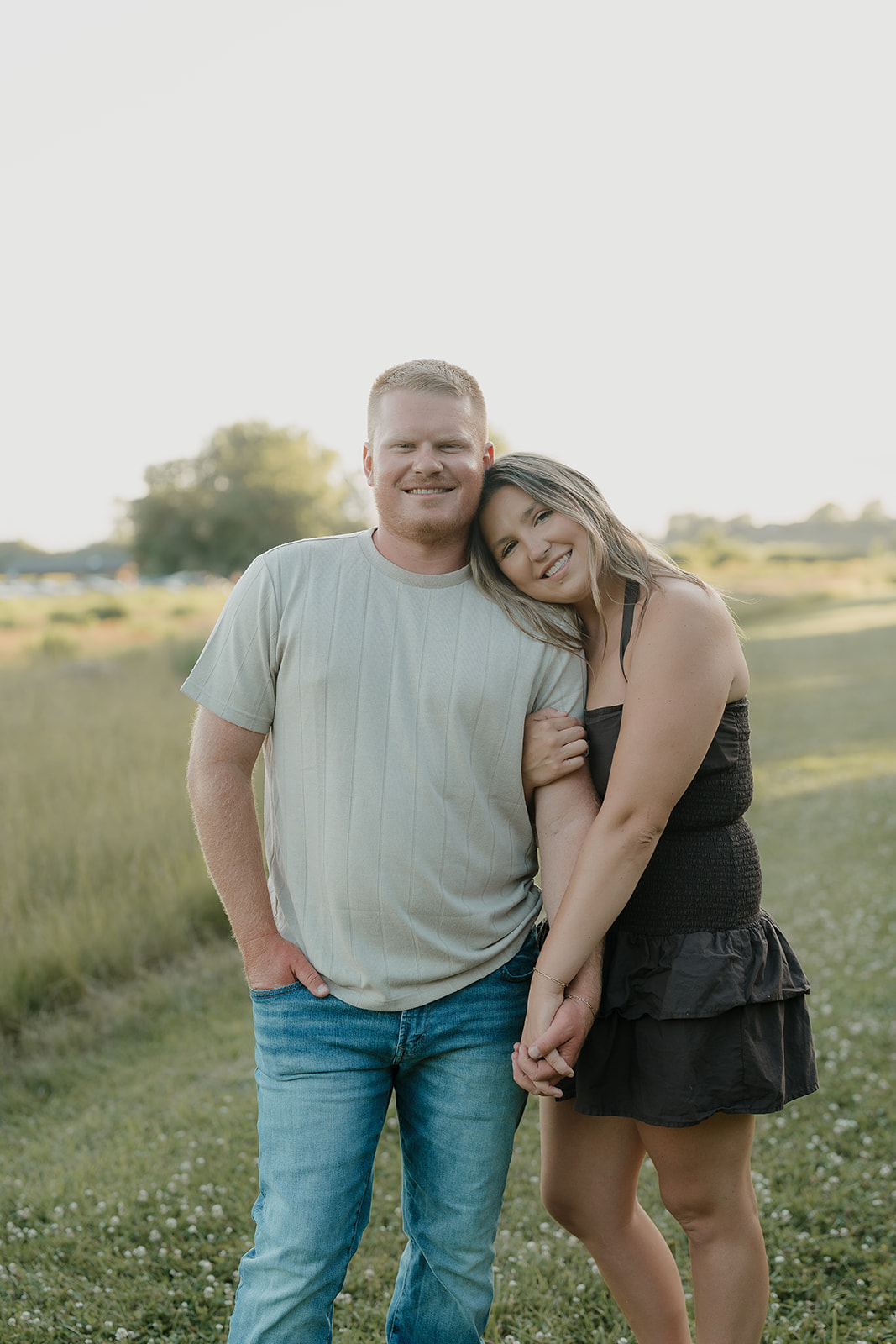 A couple stands together in a summer engagement photo outfit — dark smocked mini dress and neutral ribbed tee — leaning close and smiling in a clover field at golden hour
